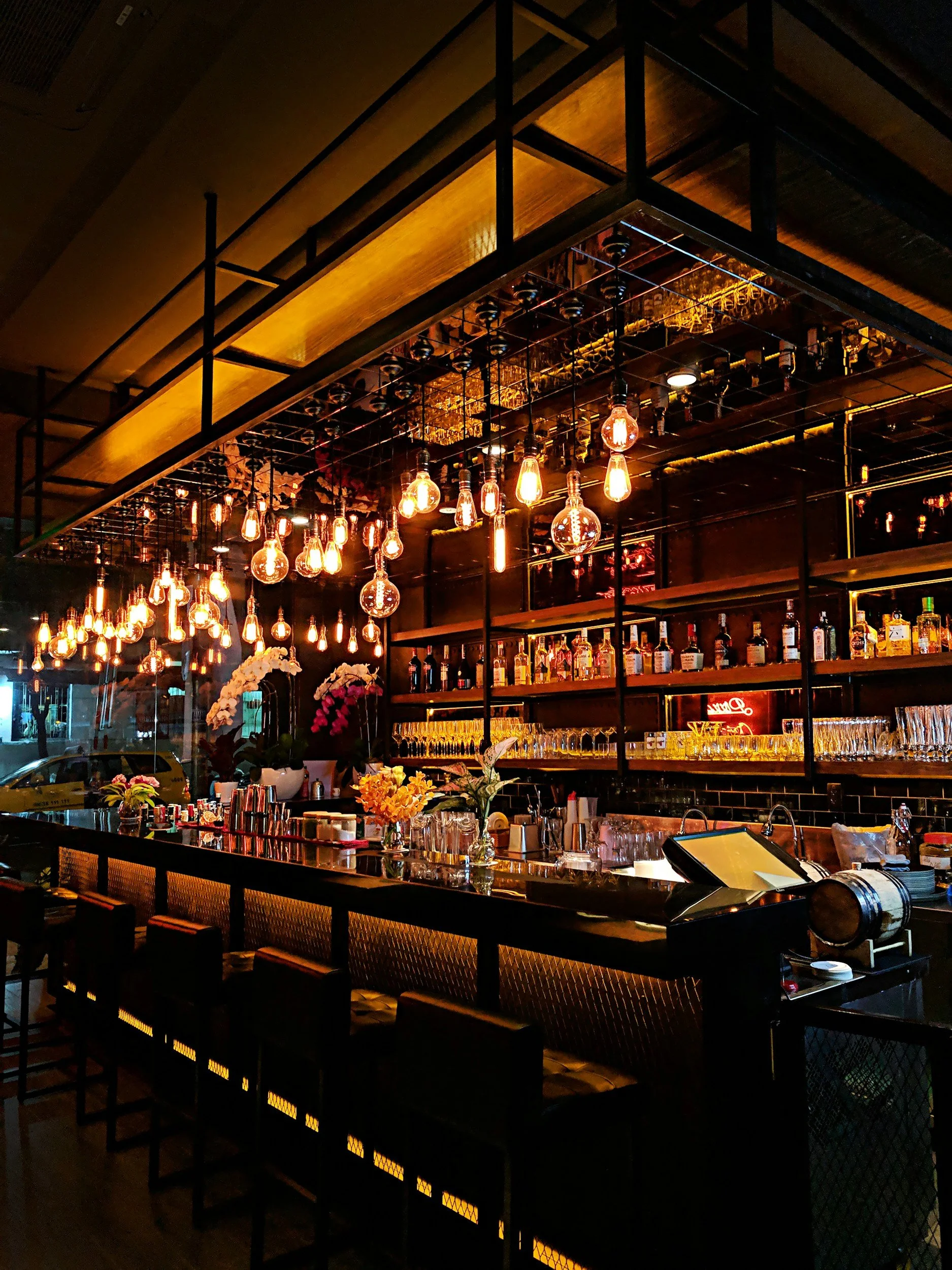A cozy bar with warm lighting, multiple hanging Edison bulbs, shelves with liquor bottles, and a floral arrangement on the counter.