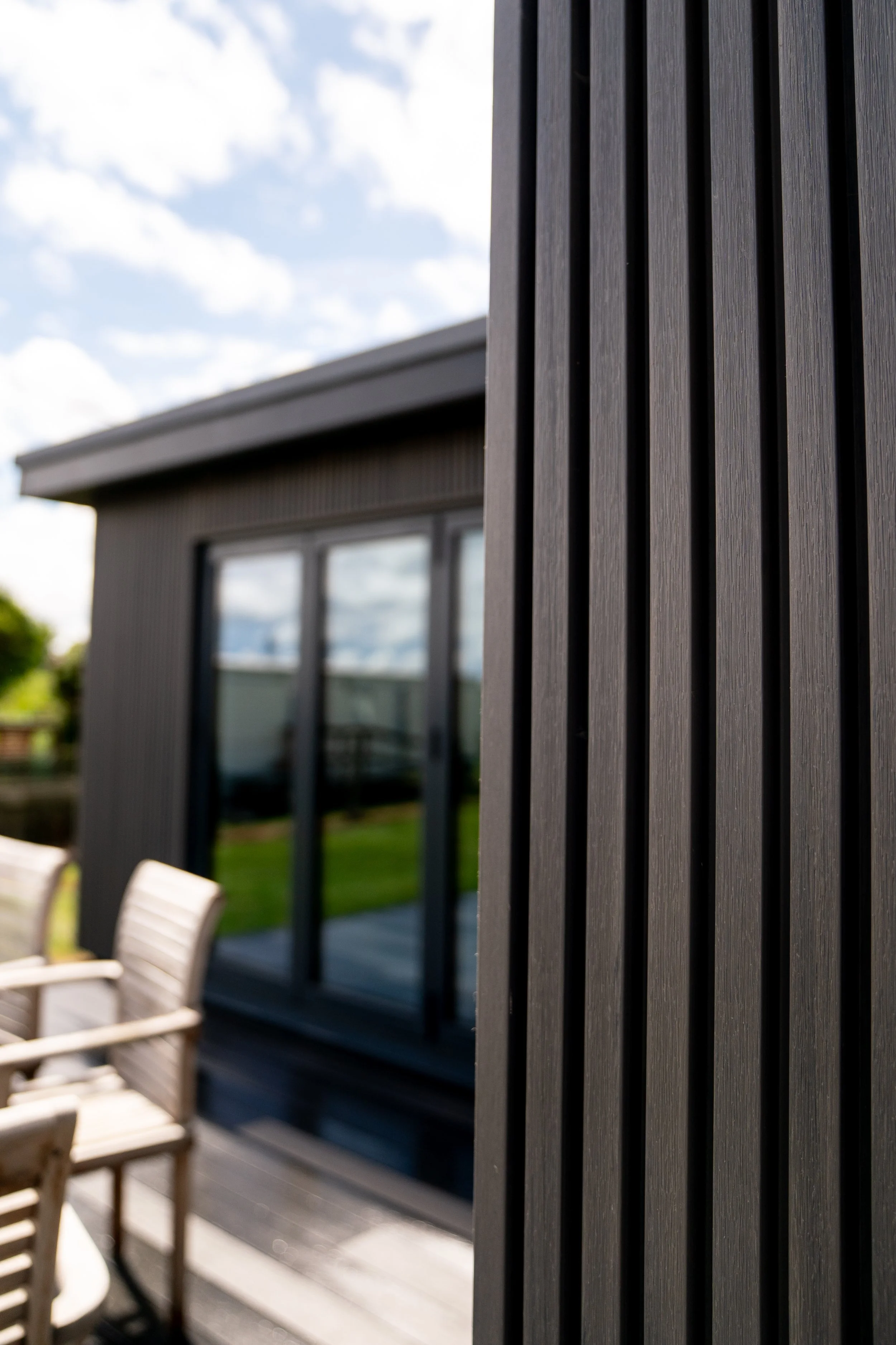 Close-up of dark outdoor wooden slats on a modern exterior wall of a house, with windows and patio furniture in the background under a partly cloudy sky.