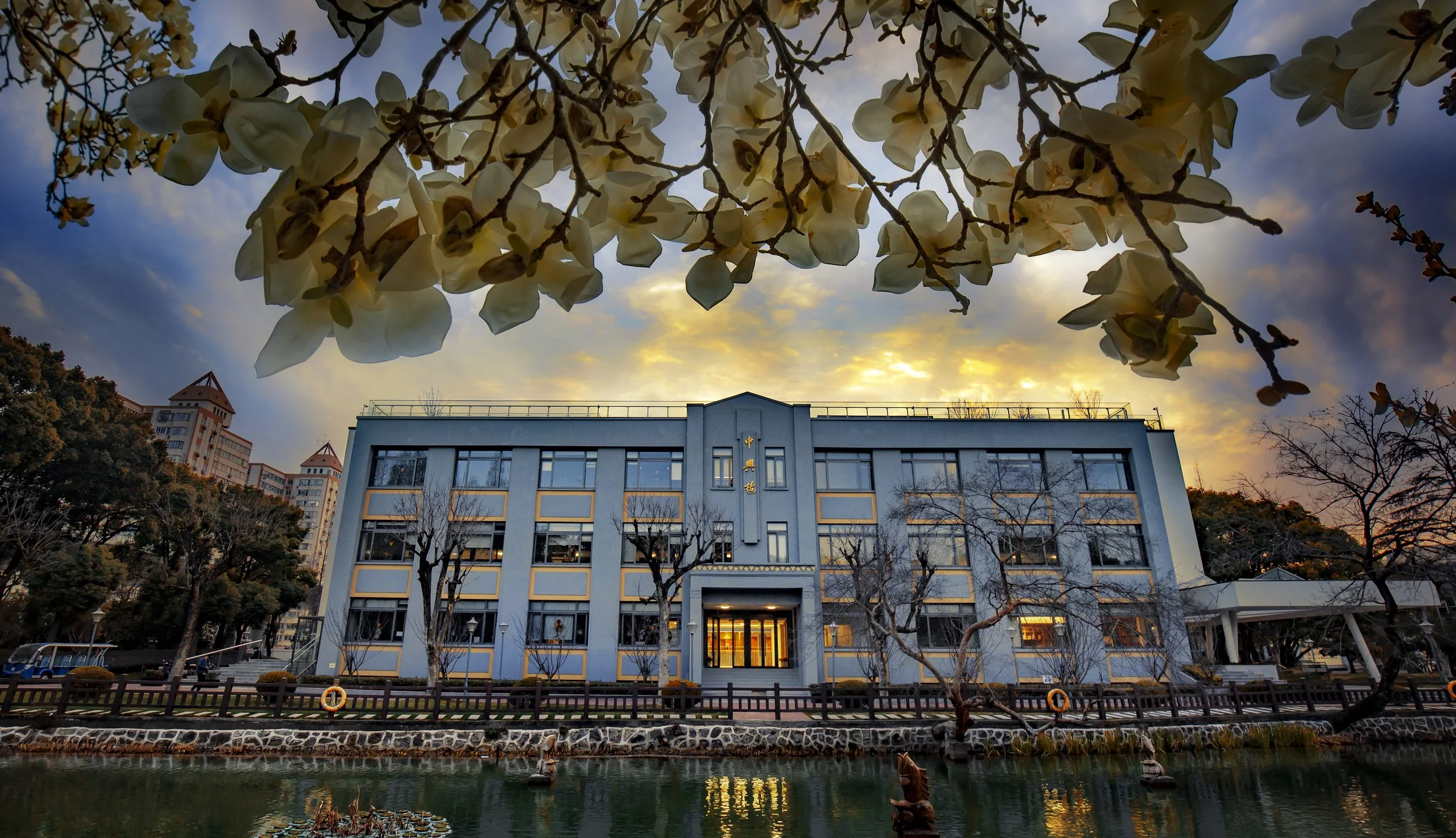 View of a classroom building with blue and white exterior, situated by a pond with ducks, under blossoming trees, during sunset with a partly cloudy sky.