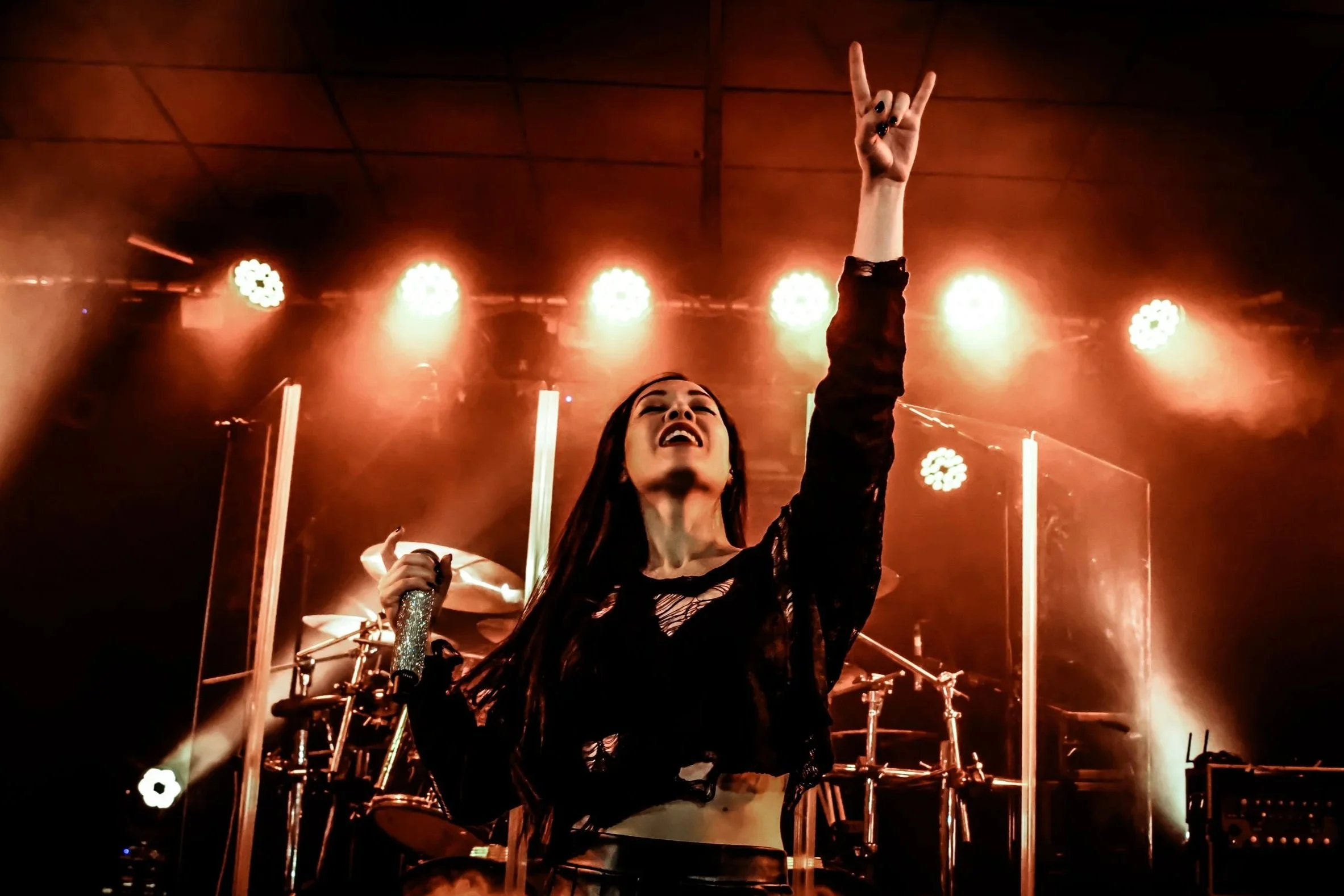 A woman on stage at a concert, raising her hand in a peace sign, with drums and stage lights in the background.