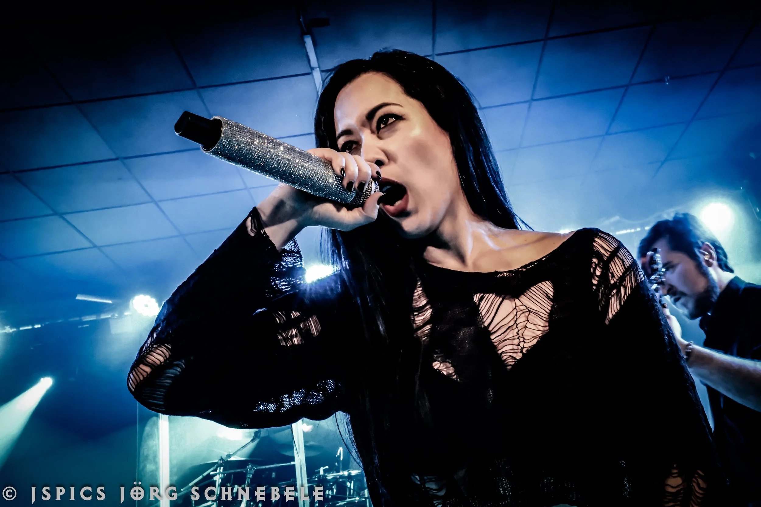 A woman singing into a microphone during a performance, wearing a black, distressed, see-through top, with a blue-toned stage and band members in the background.