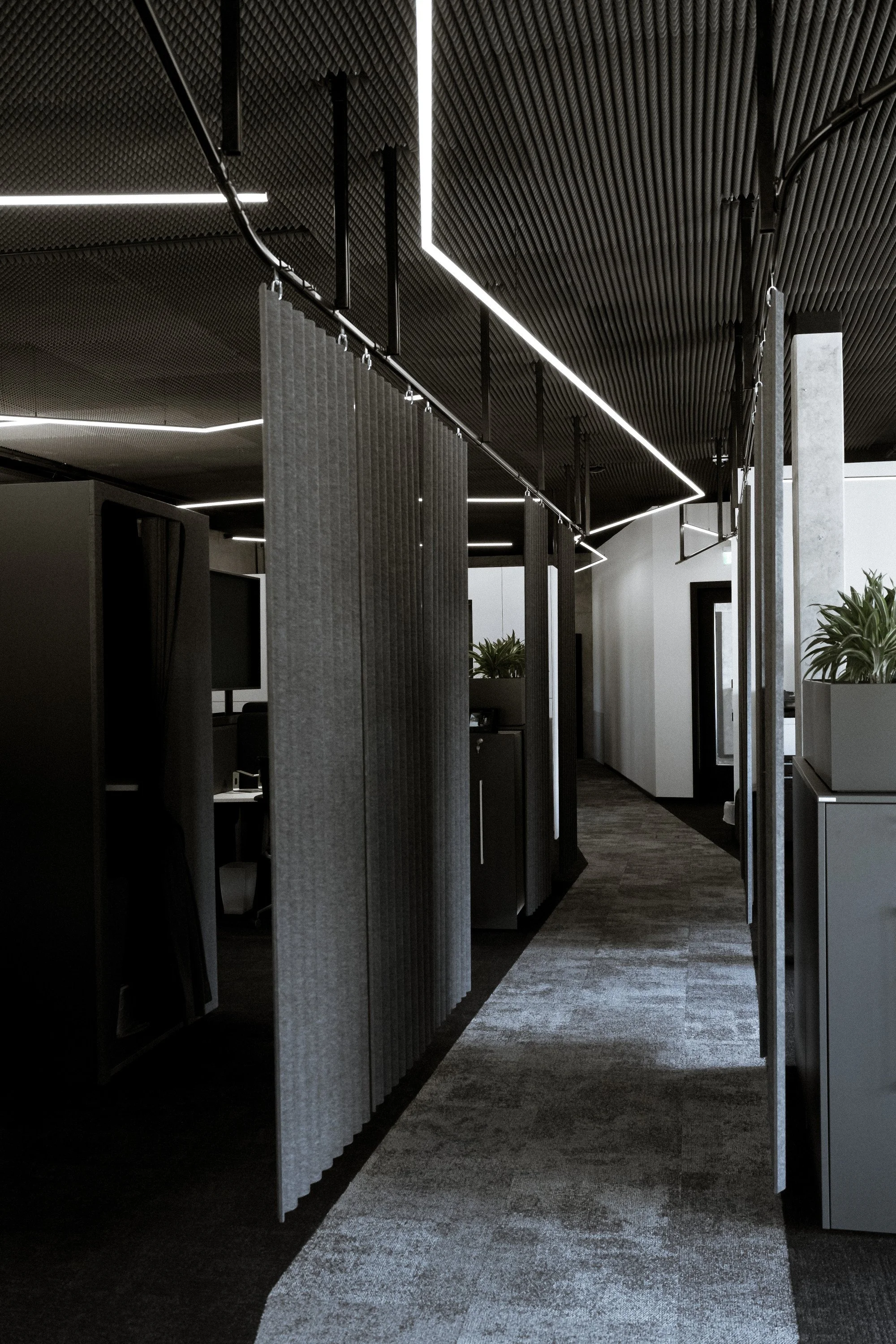 Ein moderner Büroraum mit abgetrennten Arbeitsplätzen, dunklem Deckenmaterial, LED-Beleuchtung und Pflanzen.