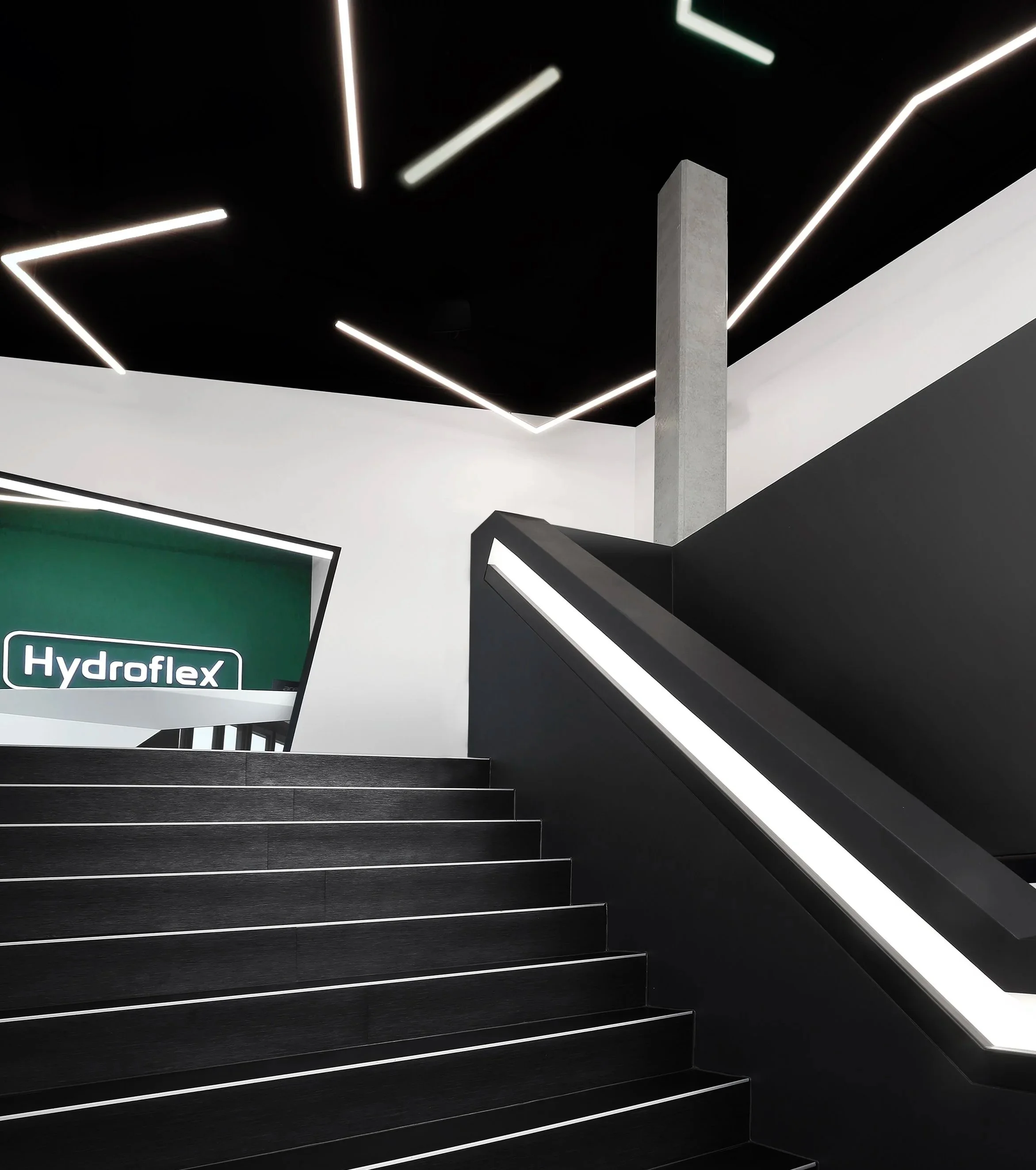 Moderne Treppe mit schwarzen Stufen, weiße Linien und eine graue Stütze, im Hintergrund eine Wand mit einem fluoreszierenden Firmenlogo 'Hydroflex'.