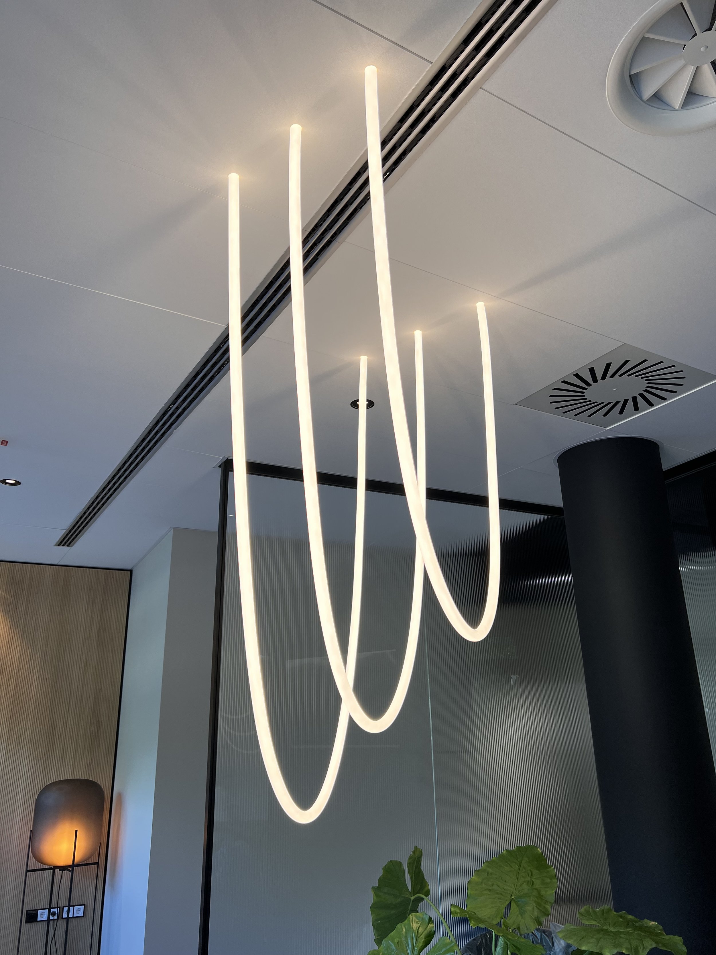 Moderne Innenraumbeleuchtung mit dekorativem, langen, weißen Neon- oder LED-Leuchtstreifen, eine Stehlampe mit Lampenschirm, eine Wand mit Steckdosen und stehendem Regal, Pflanzen im Vordergrund.