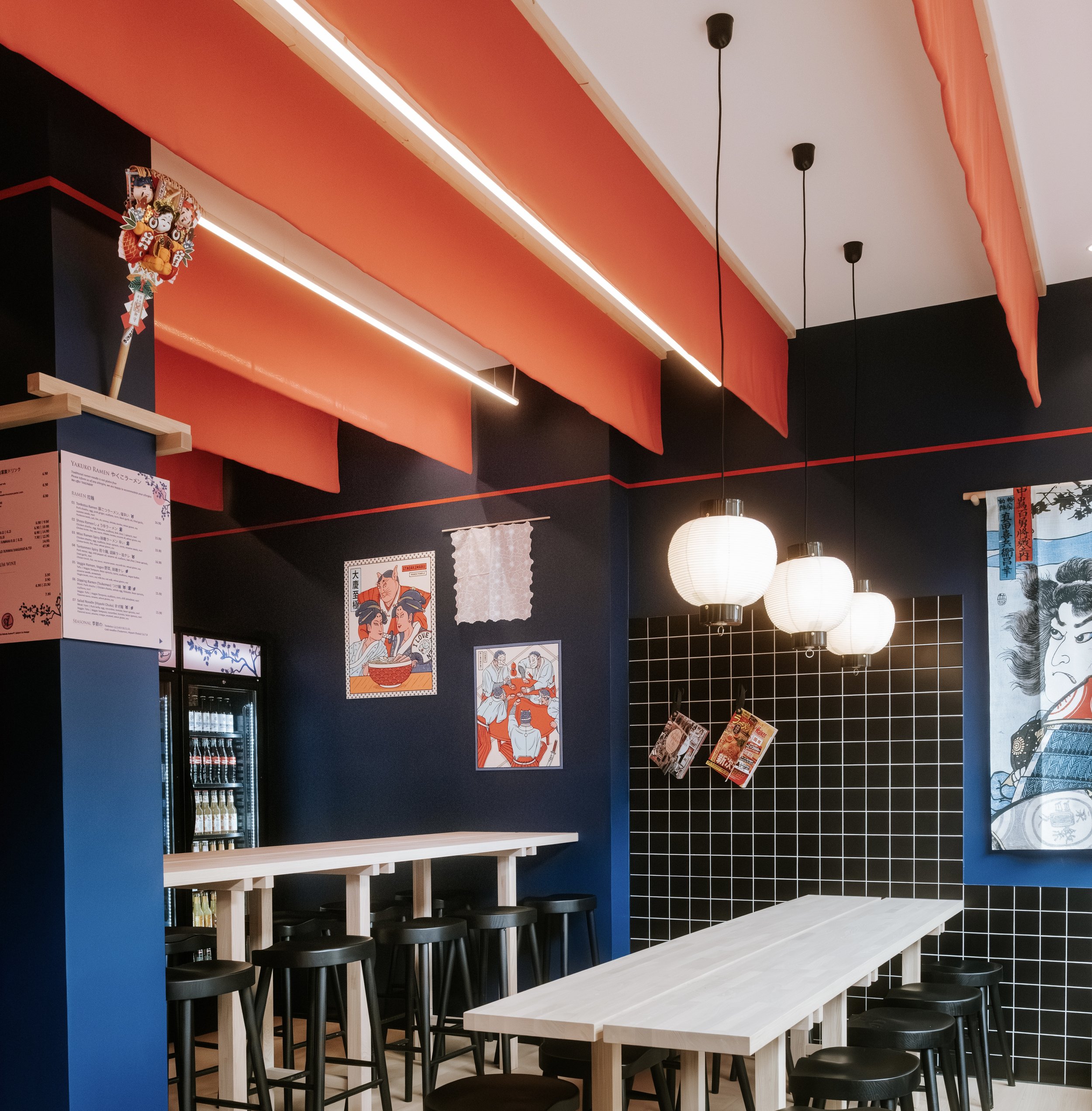 Innenaufnahme eines Restaurants mit langen Tischen, schwarzen Stühlen, Wandbildern im japanischen Stil und roten dekorativen Stoffbahnen an der Decke.