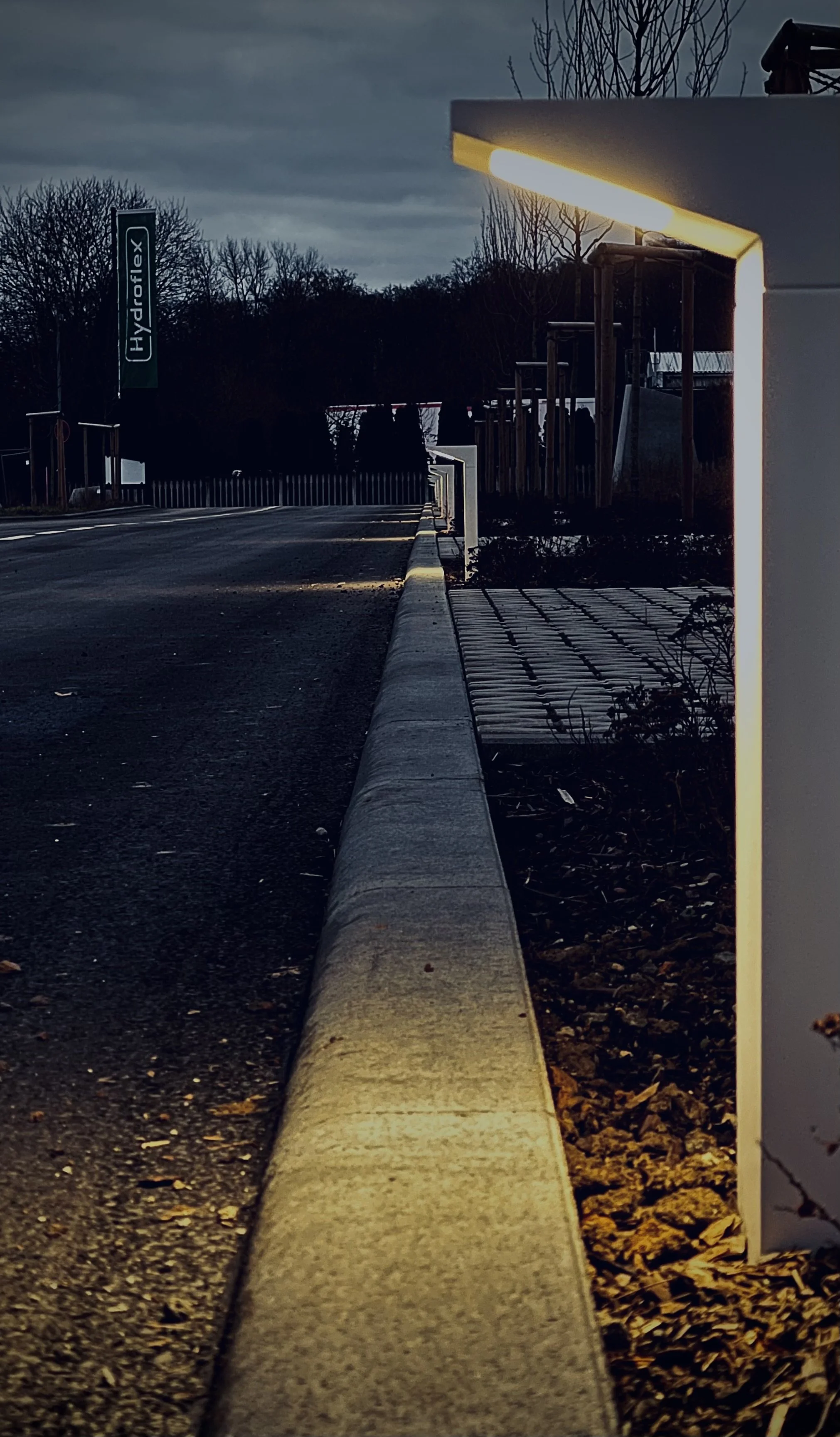 Eingeschalteter Lichtstrahler an einer Seitenwand bei Dämmerung, Blick auf eine Straße mit Gehweg und Baumrücken im Hintergrund.