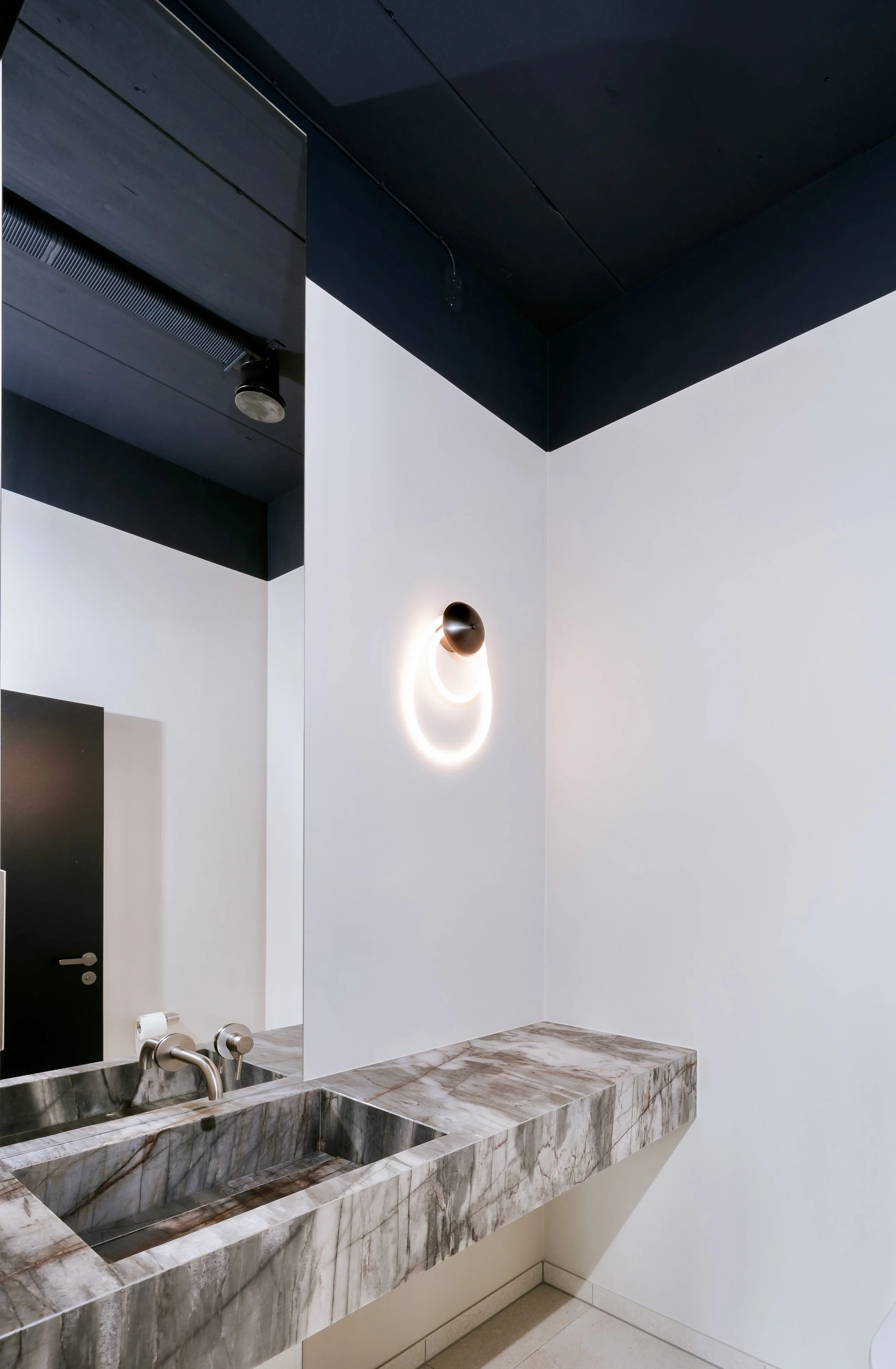 Innenansicht eines Badezimmers mit zwei Waschbecken aus Marmor, einer weißen Wand, einer schwarzen Tür, einer runden Wandlampe und einem schwarzen Deckensegment.