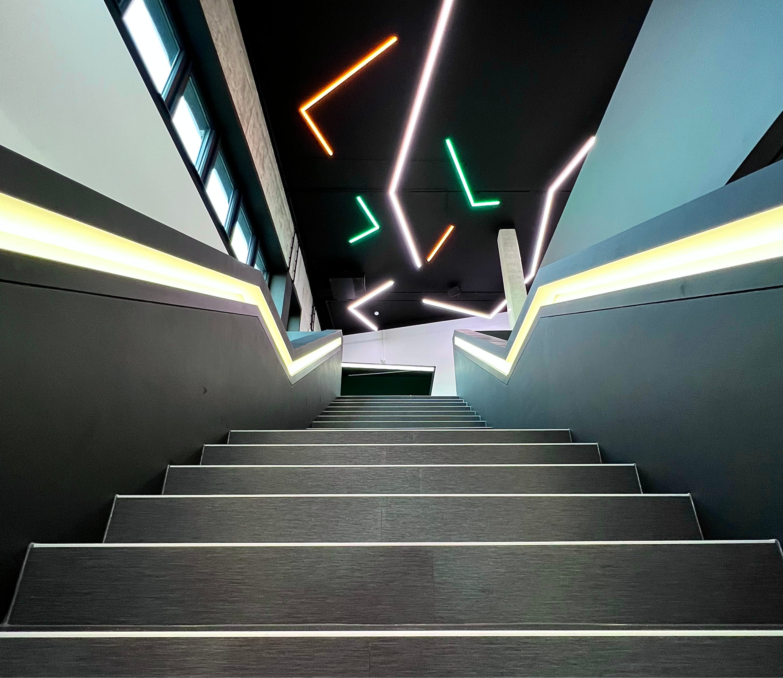 Innenaufnahme eines Treppenhauses mit schwarzen Stufen, seitlichen Beleuchtungen und einer Decke mit LED-Leuchten in verschiedenen Farben und Mustern.