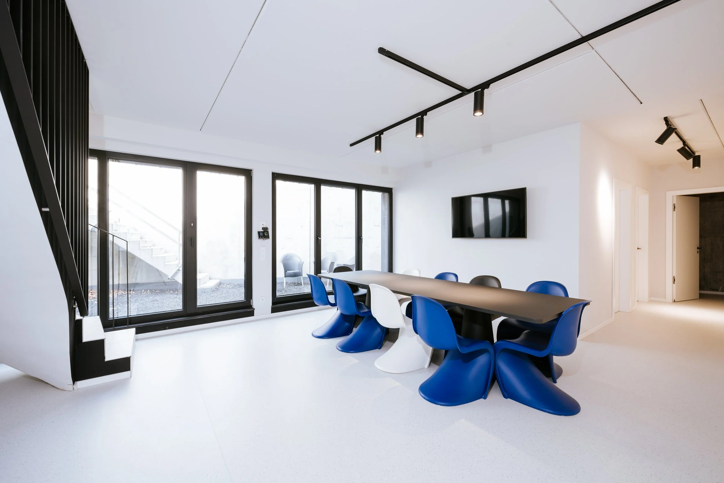 Moderner Konferenzraum mit weißen Wänden, schwarzen Fensterrahmen, einem Fernseher an der Wand, einem langen Tisch mit weißen und blauen Stühlen, großer Fensterfront und moderner Beleuchtung.