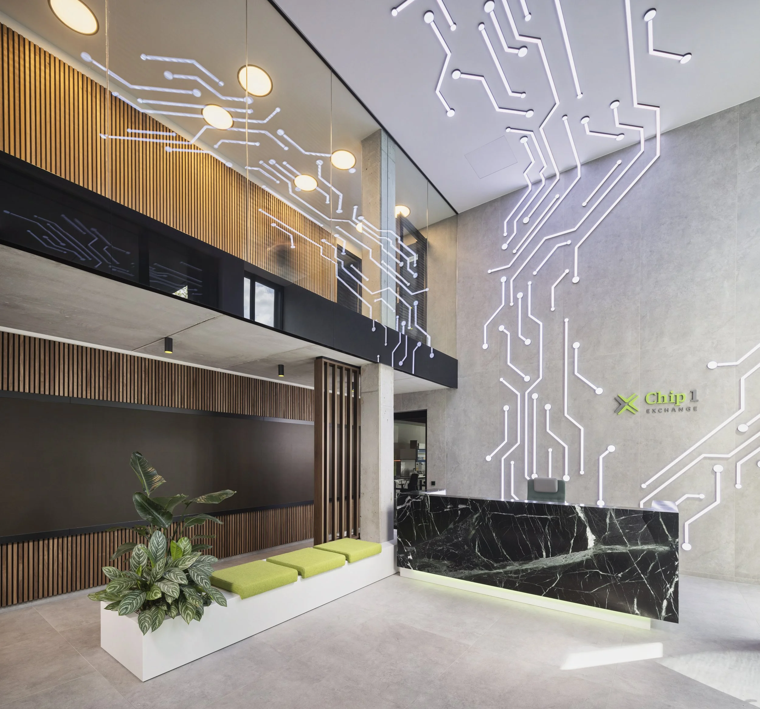 Moderne Lobby mit einer Wand aus Holzlamellen und einer weiteren Wand mit einem LED-Dekor in Form eines elektronischen Schaltkreises. Es gibt eine Betonsäule, grüne Sitzkissen, eine Pflanze und ein Empfangstresen aus schwarzer Marmor. Auf der rechten Seite befindet sich ein Logo mit dem Text 'Chip 1 Exchange'.