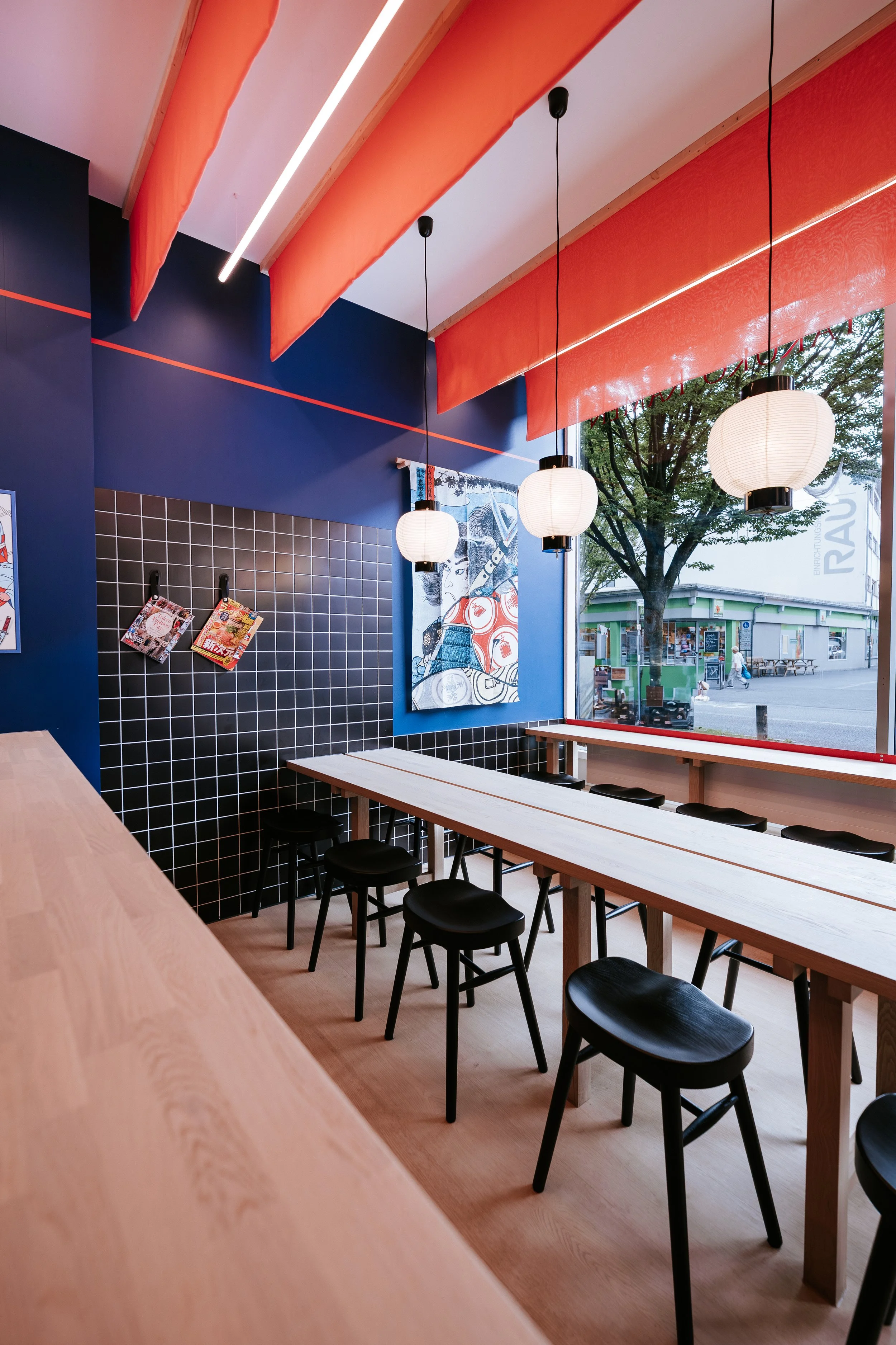 Innenansicht eines modernen Restaurants mit blauer Wand, schwarzen Hockern, einer langen Holztischreihe, weißen Papierlaternen und einer großen Fensterfront mit Blick auf eine Straße mit Bäumen und Gebäuden.