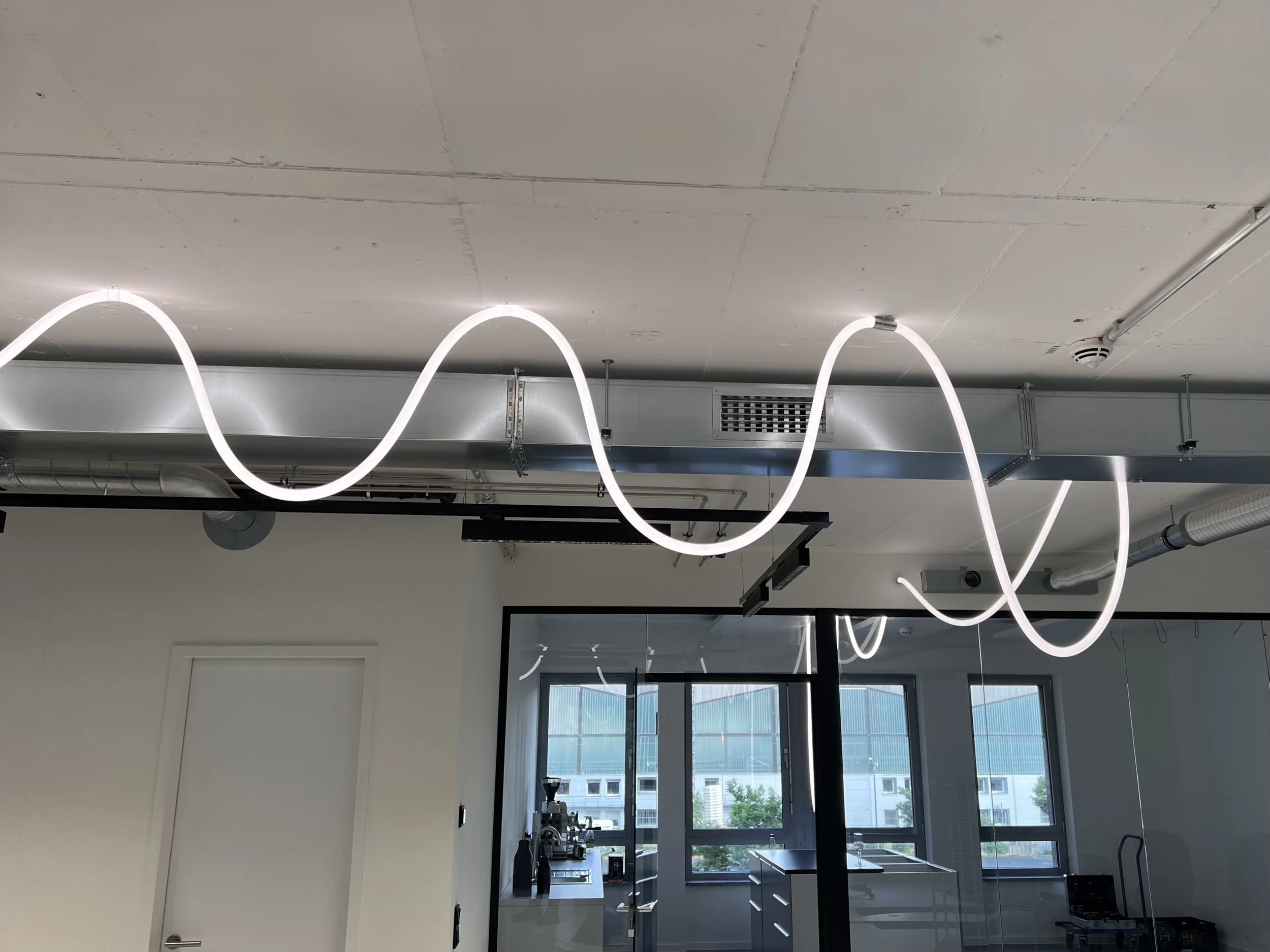 Moderner Arbeitsraum mit weißen Deckenleuchten die in geschwungenen Mustern an der Decke hängen, große Fenster mit Blick auf ein Gebäude und Tische mit technischen Geräten.