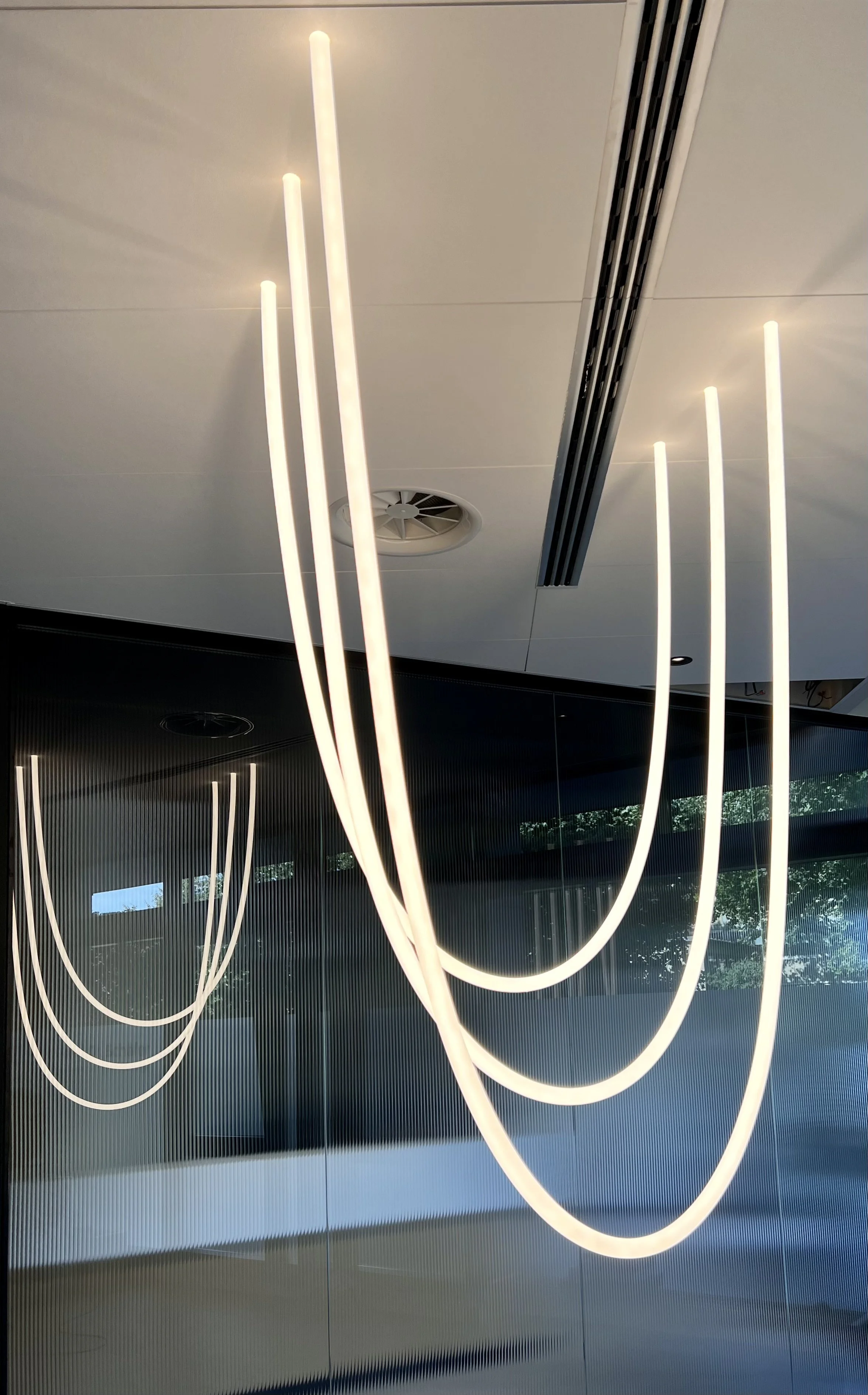 Moderne Lampe mit mehreren weißen, schlangenartigen Lichtstrahlen in einem Raum mit reflektierenden Wänden und Sicht auf Fenster mit Bäumen im Hintergrund.