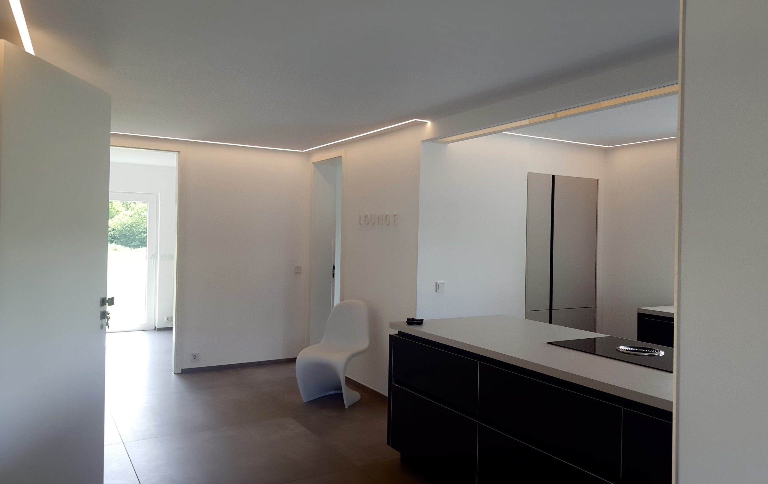 Moderner, offener Wohnzimmerbereich mit weißen Wänden, minimalistischem Design, einer weißen Stuhllehne, einem schwarzen Küchenblock, einem Kühlschrank und einer Digitaluhr an der Wand, viel natürlichem Licht durch große Fenster, mit Blick auf einen 