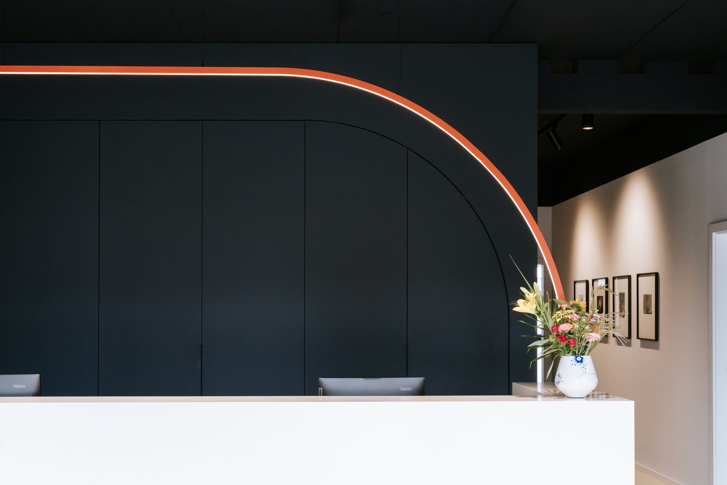 Innenraum mit dunklem Wandpaneel, einem Blumenstrauß in einer weißen Vase und einem weißen Tresen und einer Leuchtlinie.
