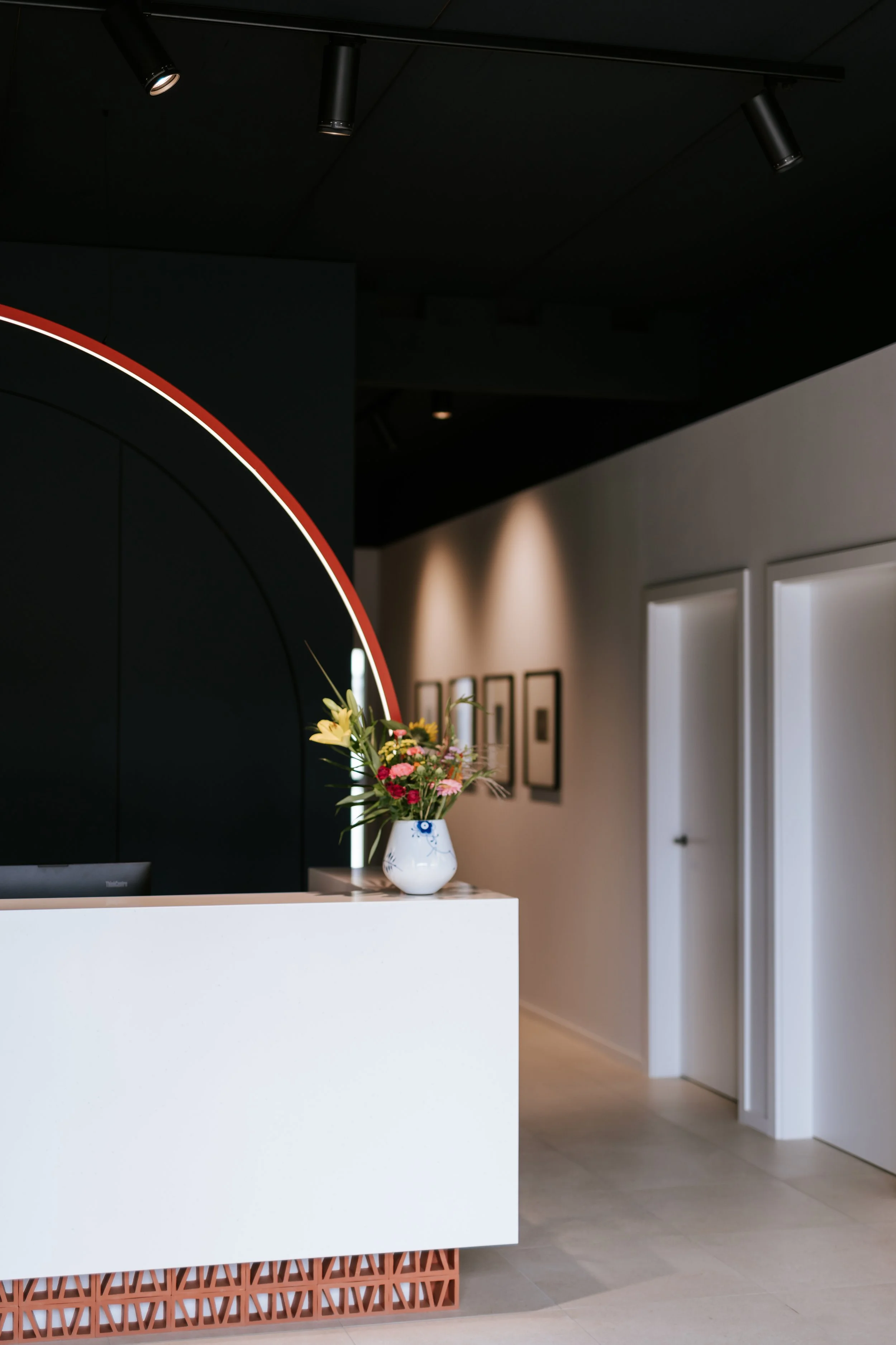 Ein weißer Empfangstresen mit einem Blumenstrauß in einer weißen Vase davor, im Hintergrund dunkle Wand mit Lichtstrahlern an der Decke.