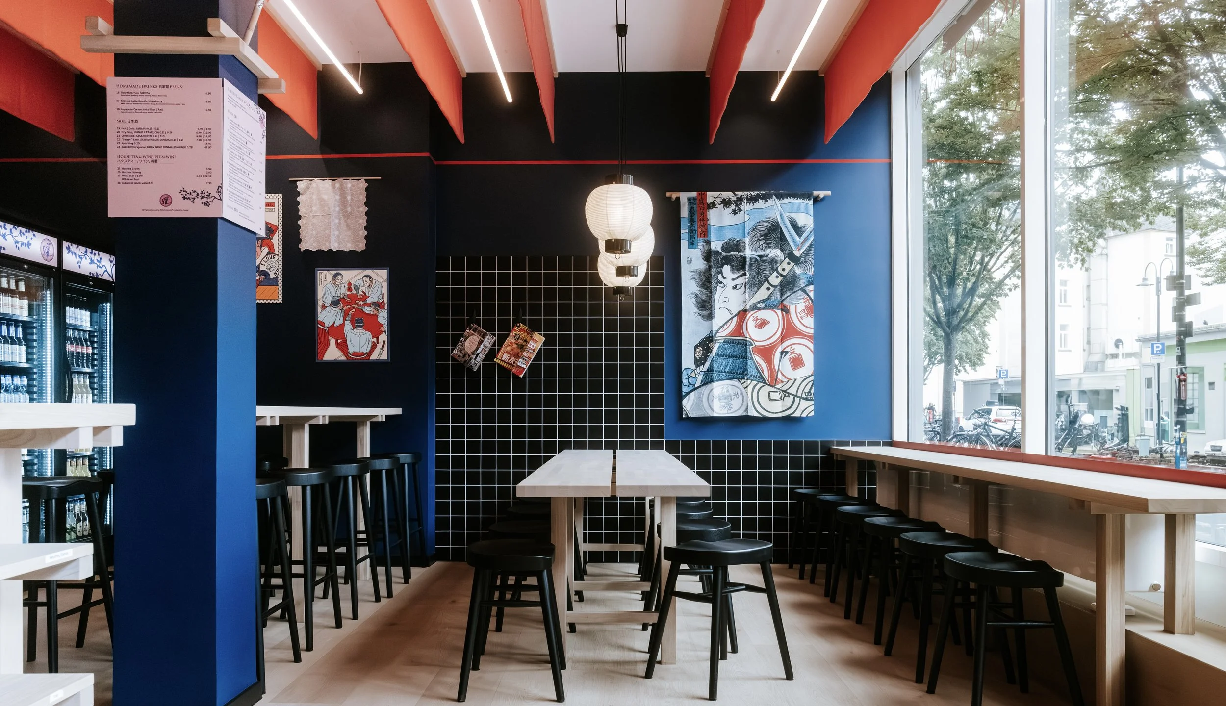 Ein gemütliches japanisch inspiriertes Restaurant mit dunklen Wänden, japanischer Kunst an der Wand, einem großen Fenster mit Blick auf die Straßen außerhalb, hängenden Lampen und Barhockern.