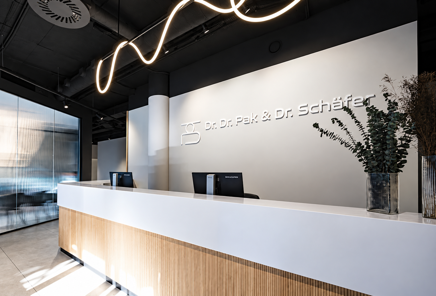 Moderne Rezeption mit weißen Theke, zwei Computer, Pflanzen in Vasen, schwarze Decke mit Leuchtstreifen, reflektierende Wand, Firmenlogo "Dr. Dr. Pak & Dr. Schäfer" an weiße Wand.