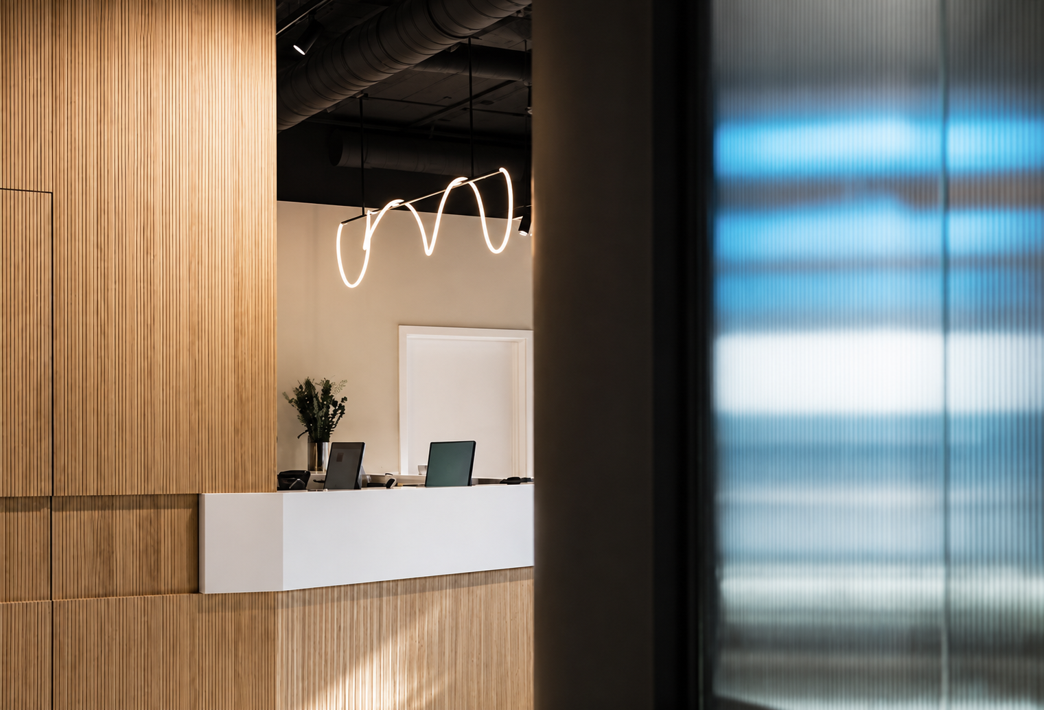 Büroempfang mit Holzverkleidung, weißer Theke, zwei Computer und einer Vase mit Grünpflanzen, moderne Beleuchtung an der Decke, Blick durch eine halbtransparente blaue Scheibe.