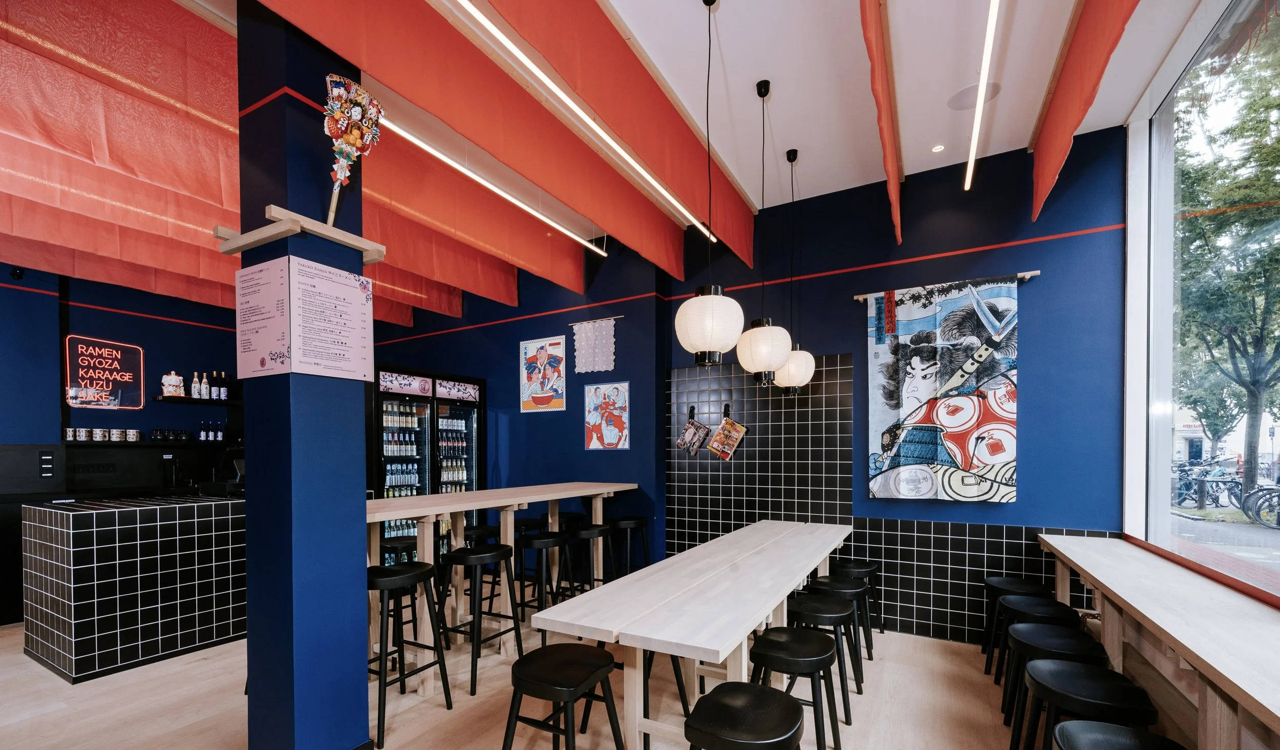 Innenraum eines modernen japanischen Restaurants mit blauer Wandfarbe, japanischer Wandkunst und rotem Stoff an der Decke, helle Holztische und schwarze Hocker.