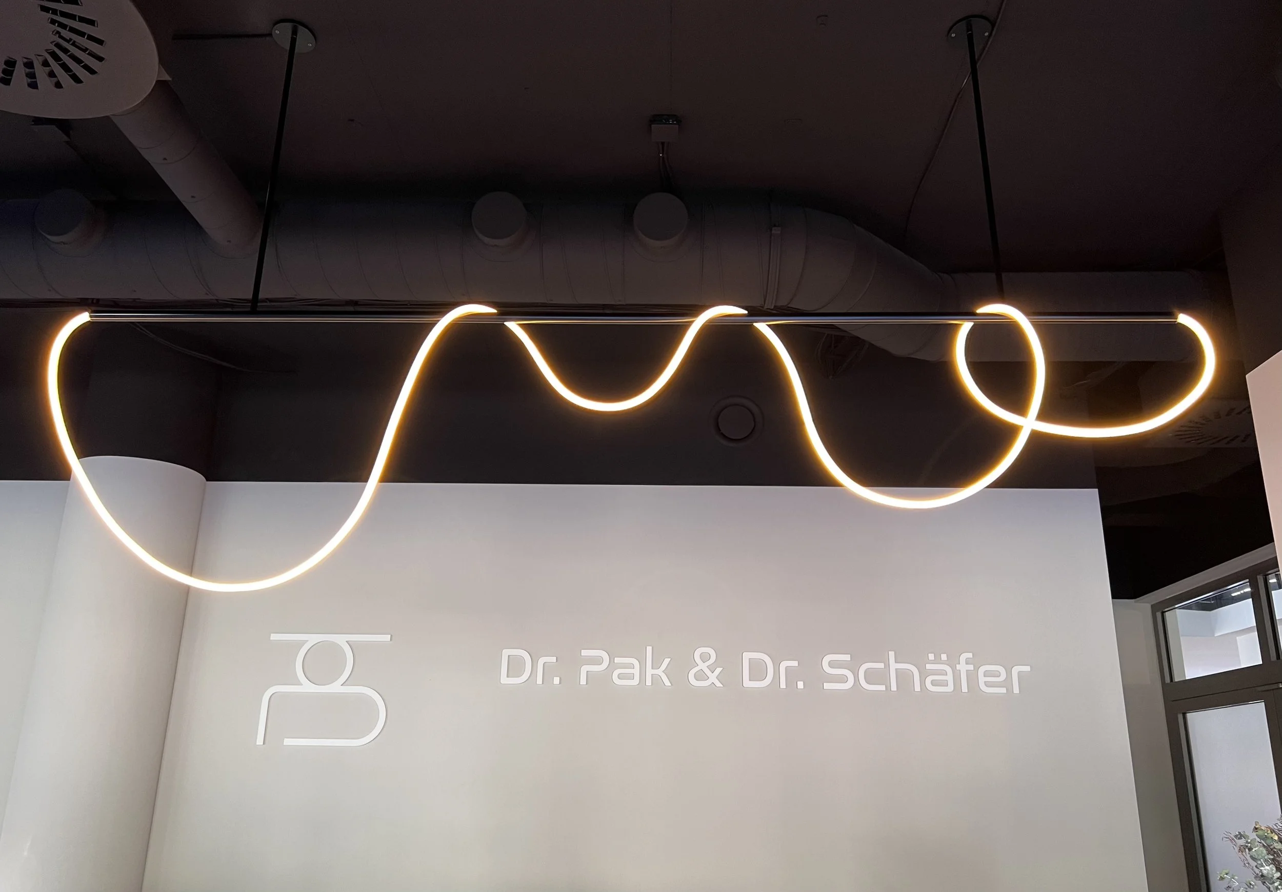 Hängende futuristische Leuchte aus Neonlicht in Form einer geschwungenen Linie, darüber Rohrleitungen an der Decke, im Hintergrund eine weiße Wand mit der Aufschrift 'Dr. Pak & Dr. Schäfer'.
