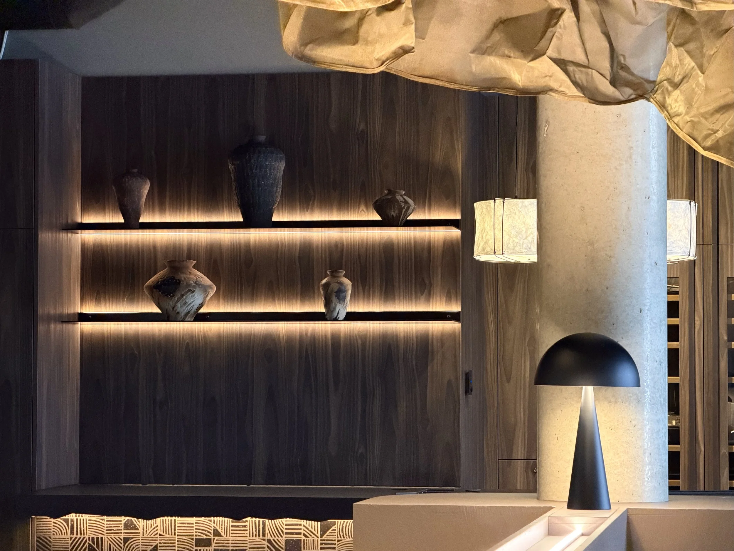 Moderner Innenraum mit dunklem Holzregal, das mehrere Keramikvasen und Leuchten enthält, sowie einem Stehlampenbesitz auf einem Steinboden im  T Kitchen in Wetzlar von Stelios Licht-Design.
