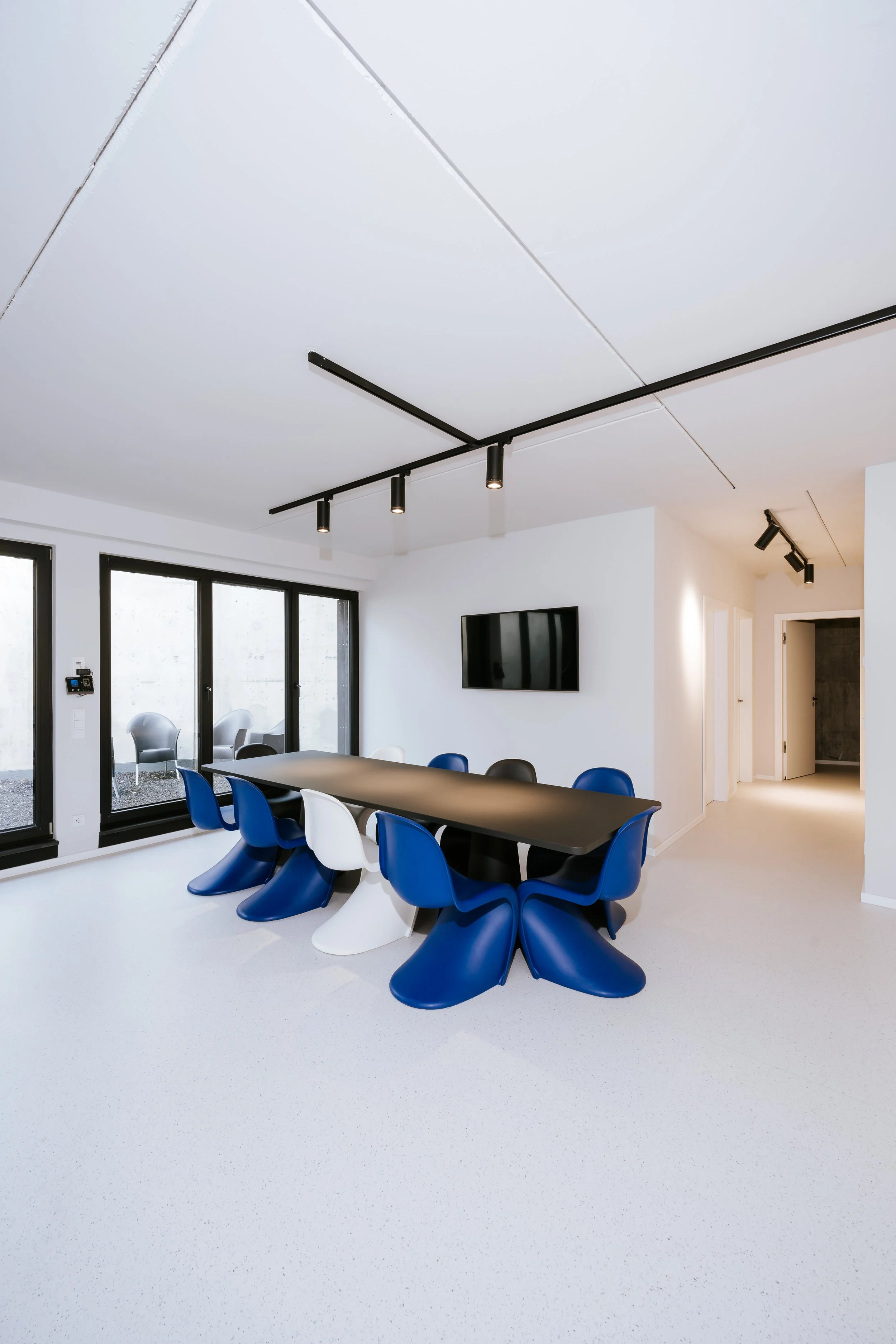 Moderner Konferenzraum mit einem schwarzen Tisch, blauen und weißen Stühlen, großen Fenstern, einer Wand mit einem Flachbildfernseher und moderner Beleuchtung.