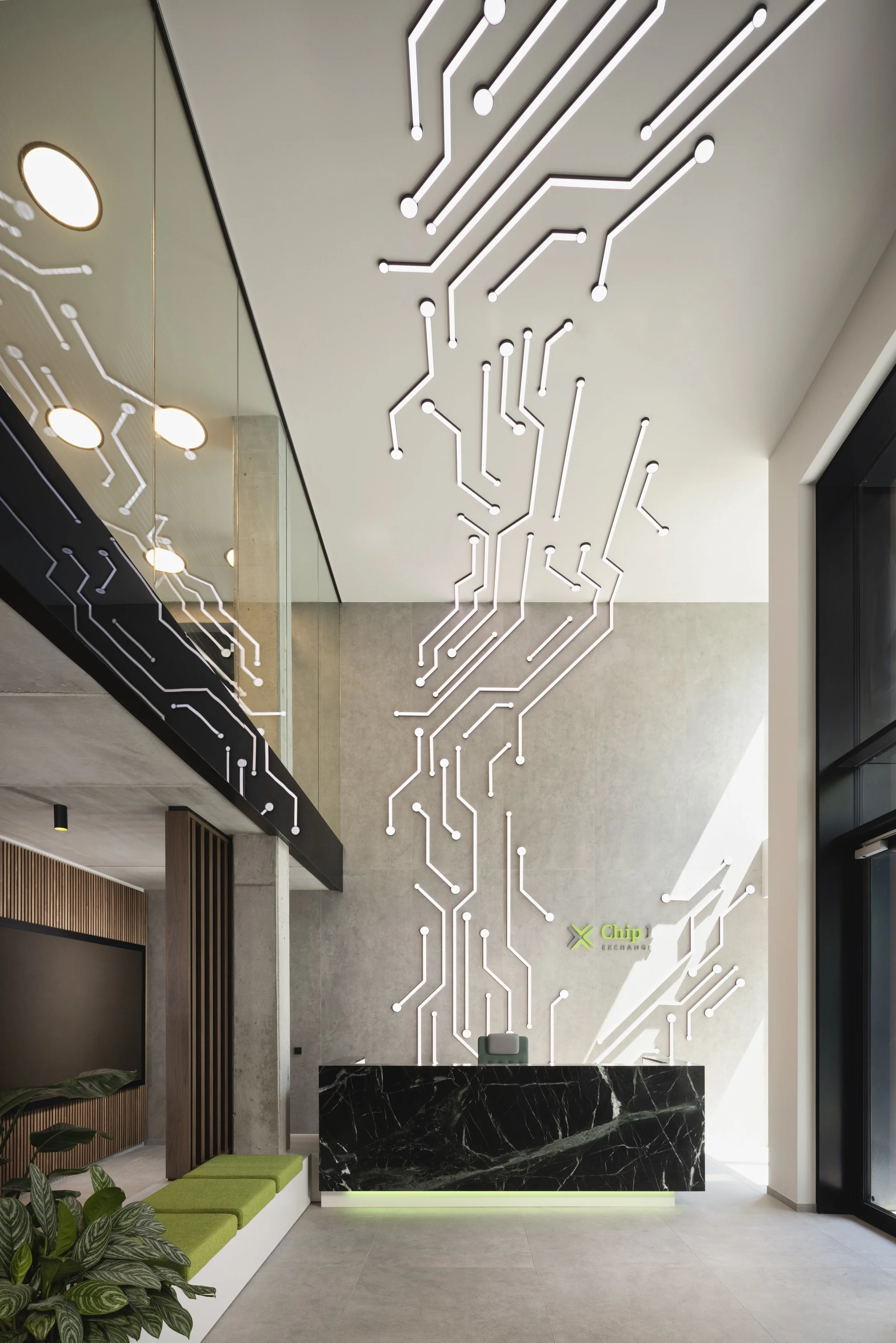 Modernes Empfangsbereich mit Wanddekoration in Form eines weißen LED Schaltkreis-Musters und dem Logo 'Chip 1 Exchange' auf der Wand, schwarzer Marmortresen und Sitzbank aus Grüntönen. Es gibt Pflanzen und eine Treppe mit Glas- und Holzgeländer.