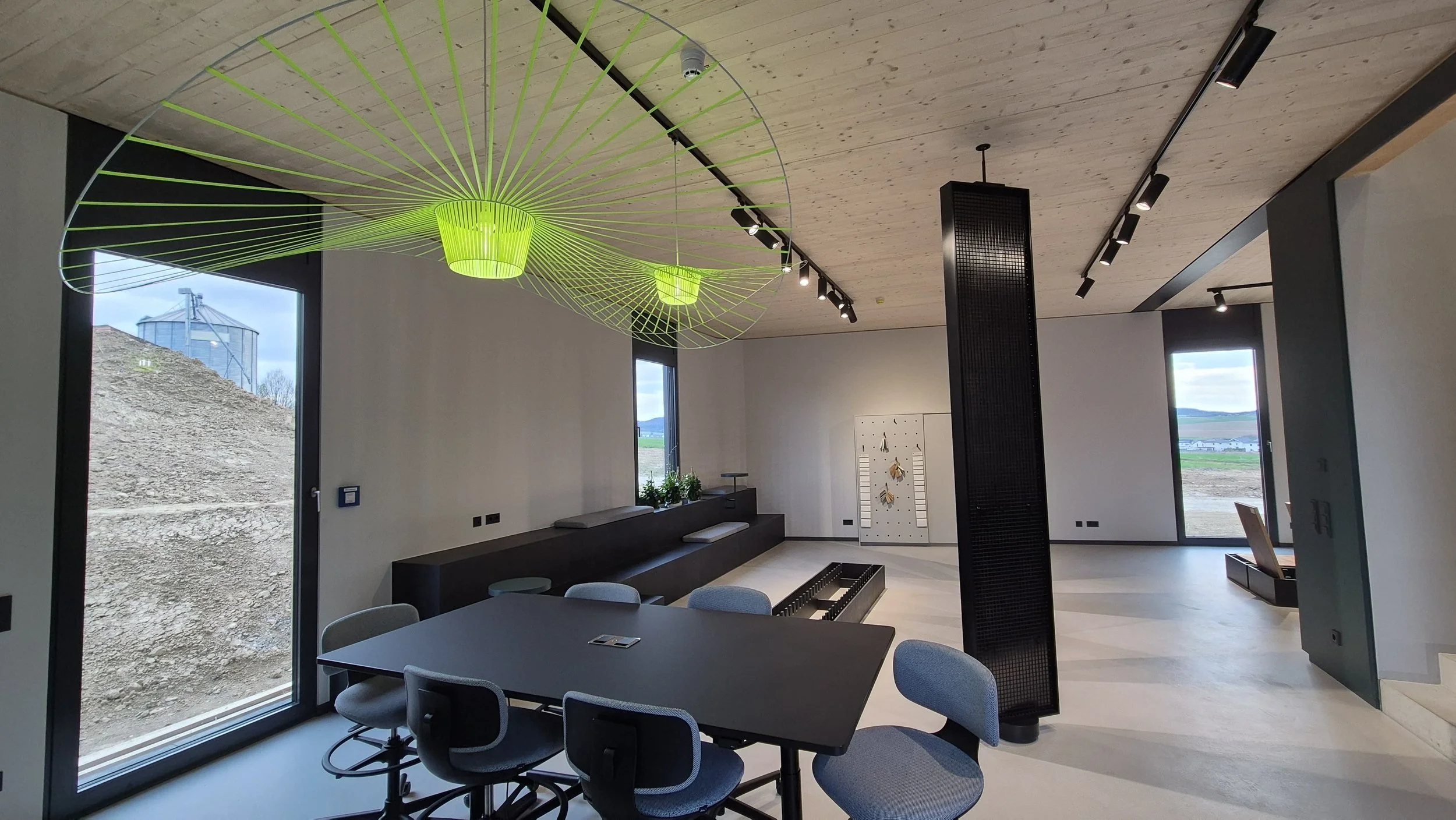 Moderner Raum mit einem runden Tisch, Stühlen, großen Fenstern, einer Holzdecke und einer modernen Lichtinstallation in leuchtendem Grün. Es gibt Blick nach draußen auf eine ländliche Landschaft.