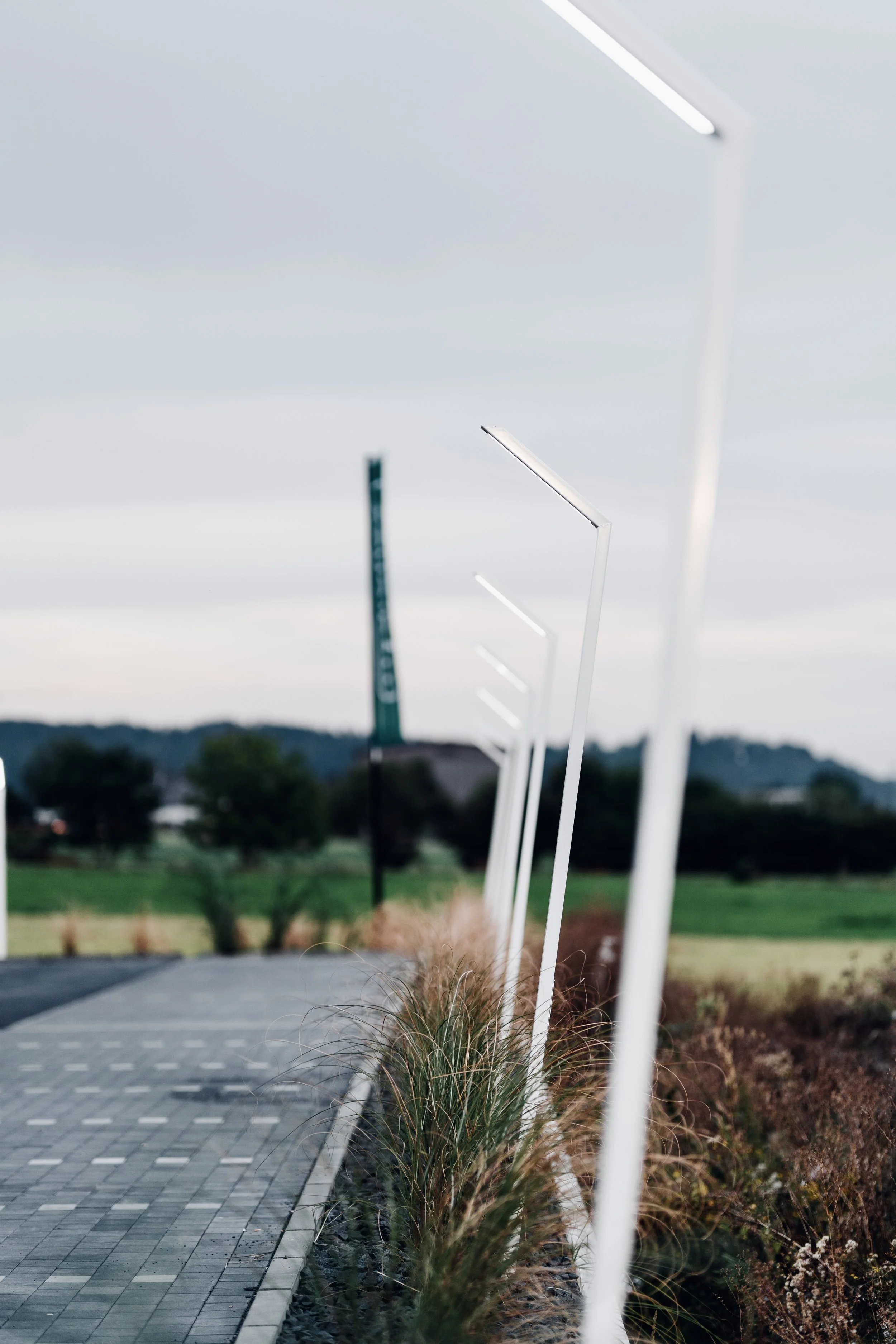 Leere Parkplätze mit weißen Solarleuchten entlang eines Gehwegs, im Hintergrund Bäume und bewölkter Himmel.