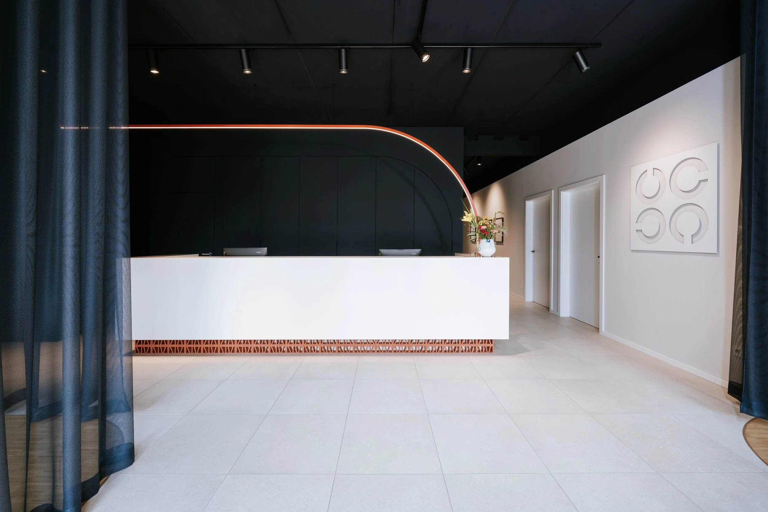 Moderne Empfangstheke in einem hellen Raum mit dunkler Decke, weißen Wänden und dekorativen Kunstwerken, ergänzt durch einen Blumenstrauß in einer weißen Vase. LED Beleuchtung elegant integriert in den Bogen über der Rezeption.
