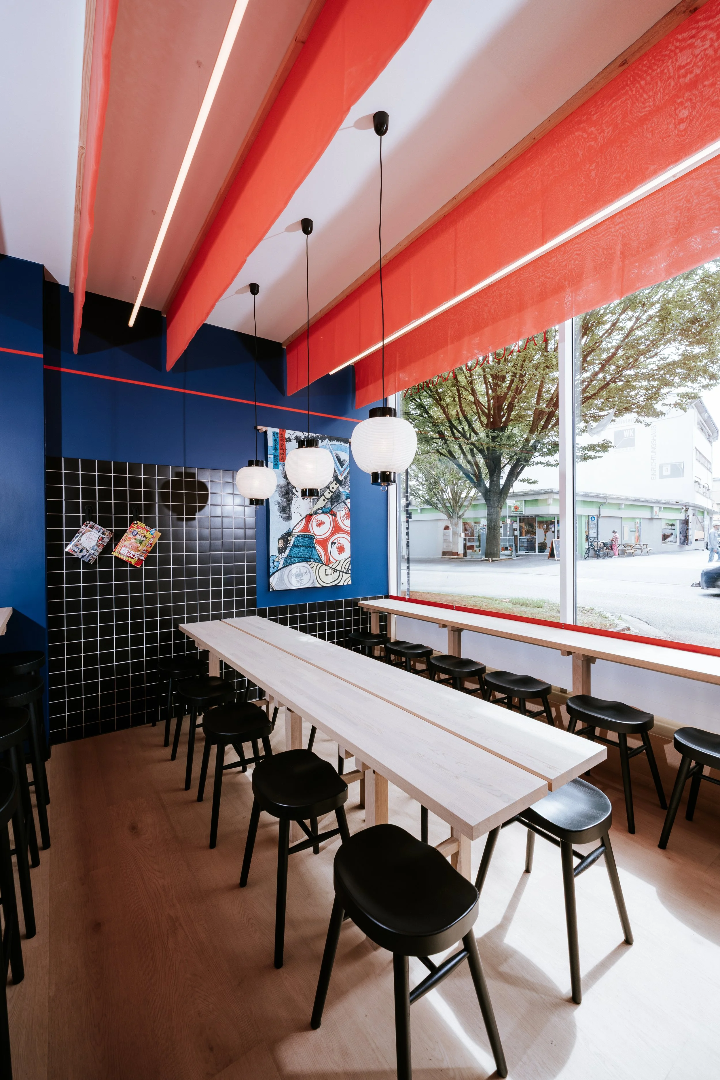 Ein modernes Café mit langen weißen Tischen, schwarzen Hockern und dunkler Wanddeko. Große Fenster lassen viel Tageslicht herein, Blick auf einen Baum und die Straße draußen.
