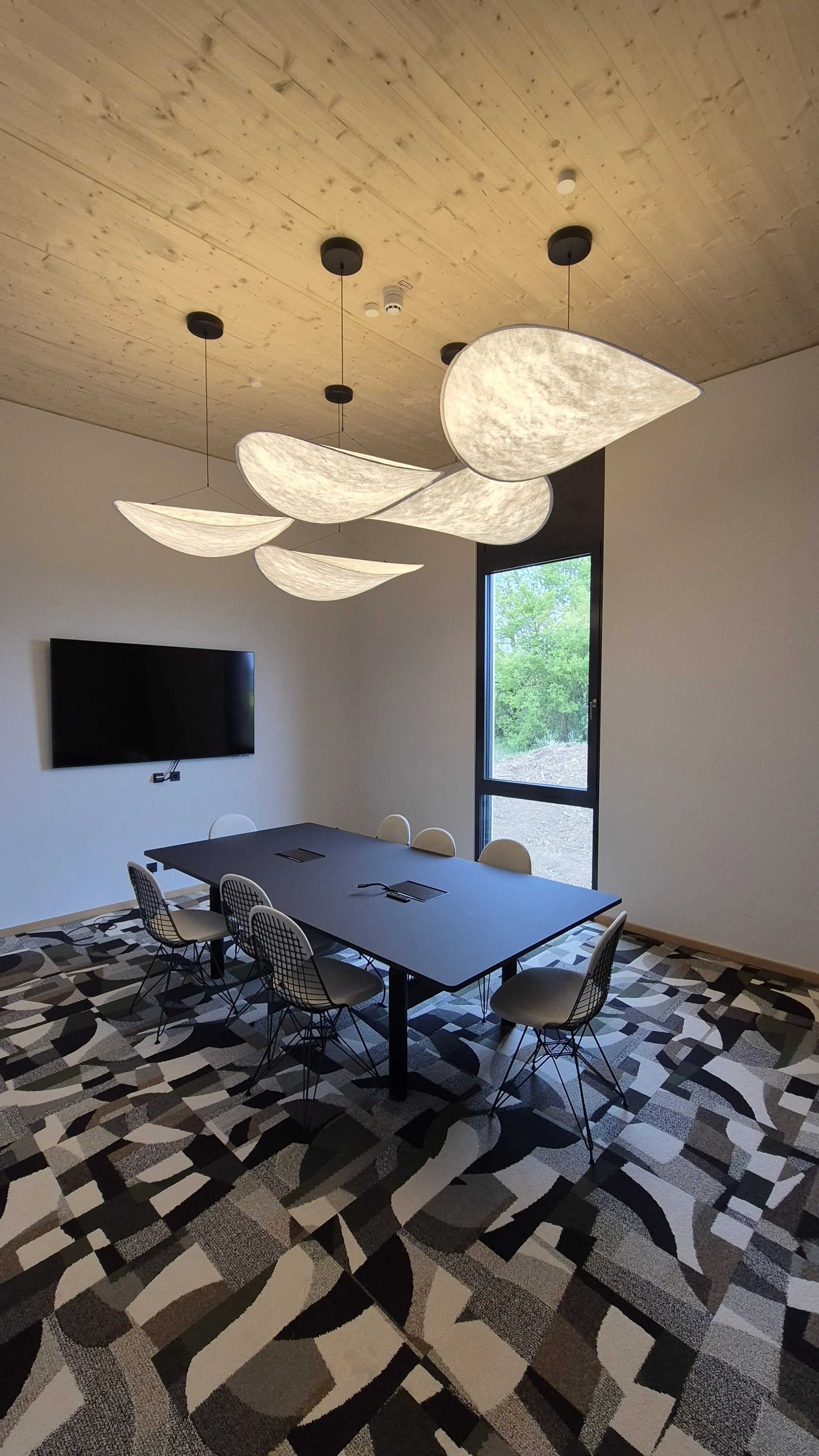Moderner Konferenzraum mit schwarzen Tisch, weißen und schwarzen Stühlen, Teppichboden mit geometrischem Muster, Wandraum mit schwarzem Fernseher und großes Fenster mit Blick nach draußen, Deckenbeleuchtung mit weißen und schwarzen Lampenschirmen