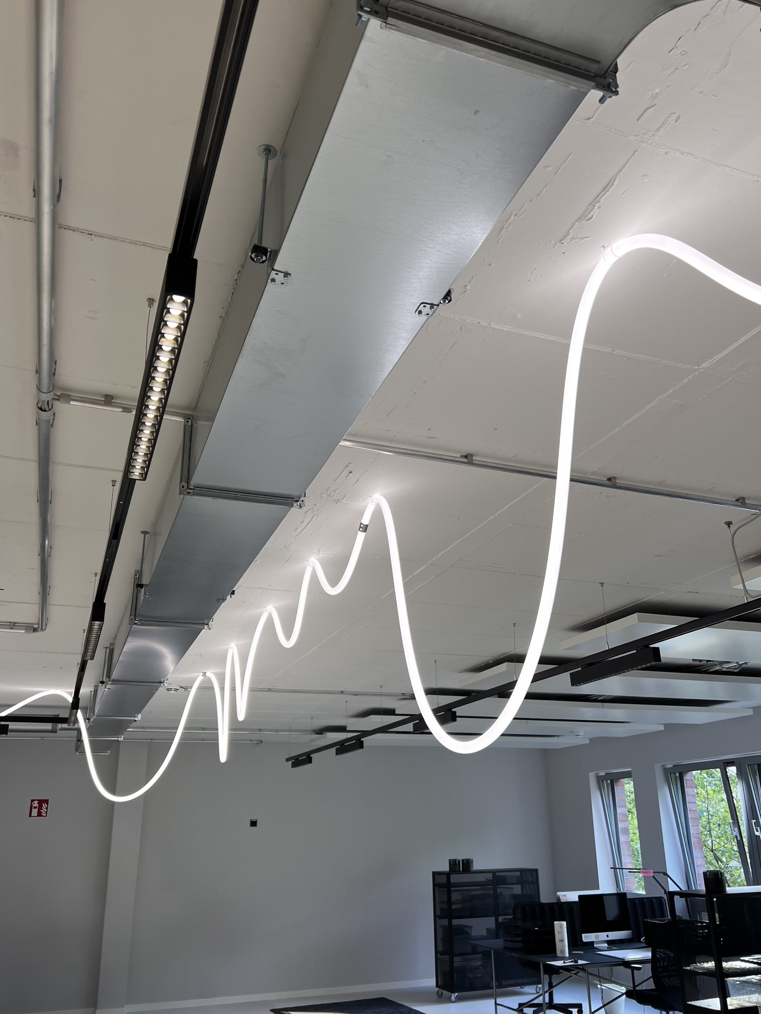 Moderne Büroetage mit weißen Decken, einer Wellenförmigen LED-Lichtleiste, großen Fenstern mit Blick nach draußen, und mehreren schwarzen Büchertischen und Stühlen.
