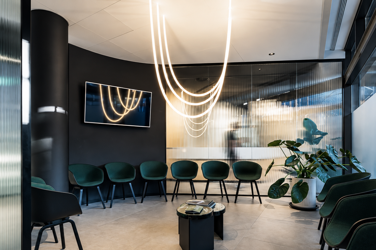 Moderner Wartebereich mit grünen Stühlen, einer schwarzen runden Tischfläche mit Zeitschriften und einem Fernseher an der Wand, beleuchtet durch neonartige 360 Grad Lichtketten, mit einer großen Zimmerpflanze.