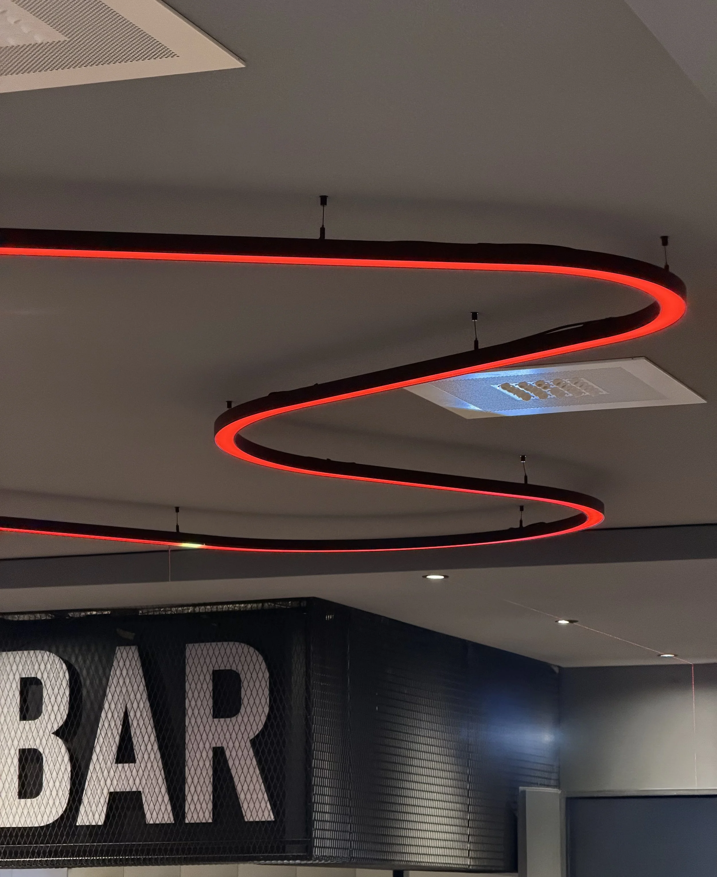 Moderne Deckenbeleuchtung mit spiralförmigen roten Lichtbändern in einem Innenraum und einem Mesh-Design mit großen Buchstaben im Hintergrund im Dom Palais Wetzlar von Stelios Licht-Design.