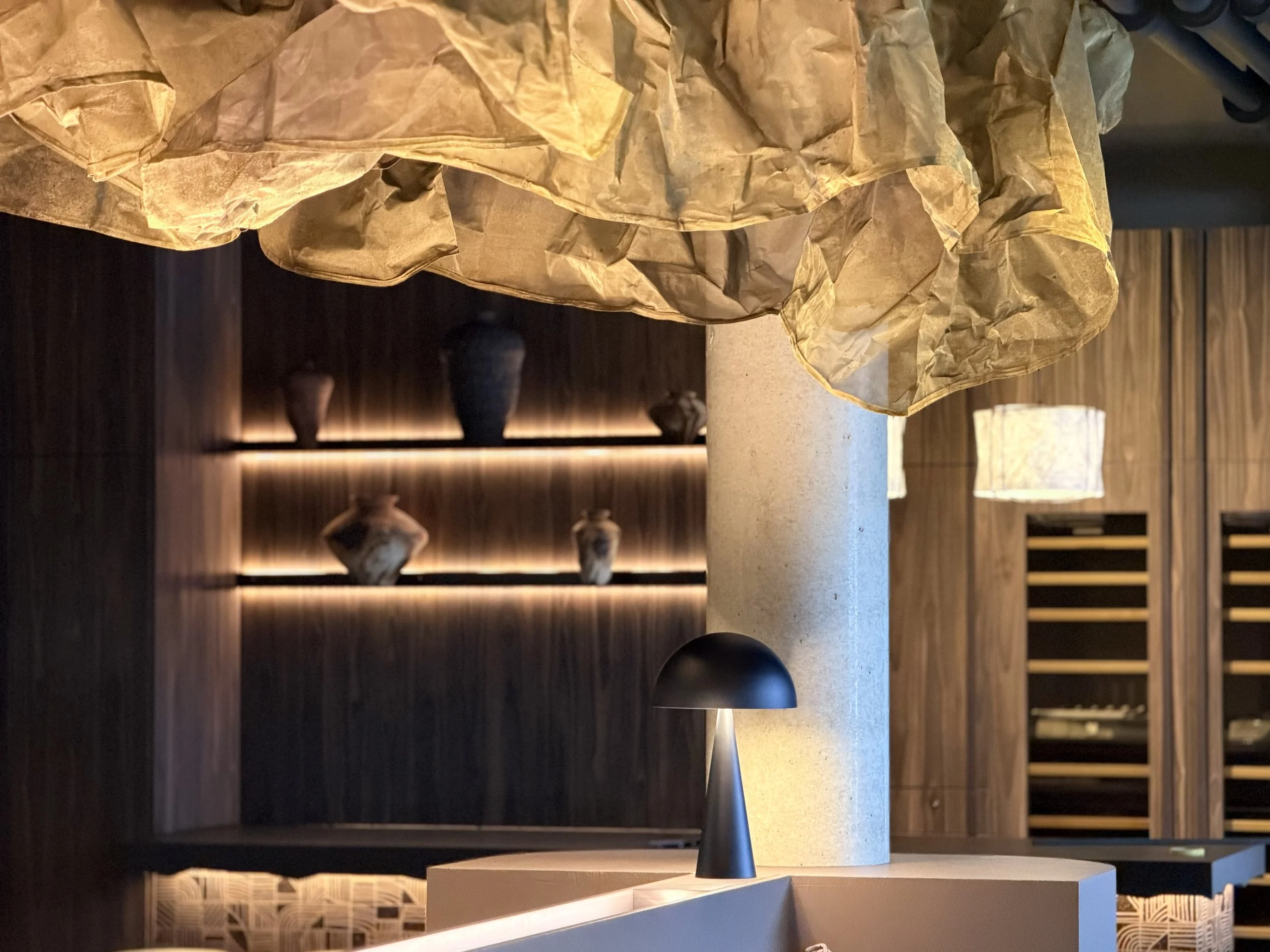 Ein moderner Tischlampe mit schwarzem Lampenschirm auf einem weißen Tisch, im Hintergrund eine Holzregal mit Dekorationsgegenständen im T Kitchen in Wetzlar von Stelios Licht-Design.