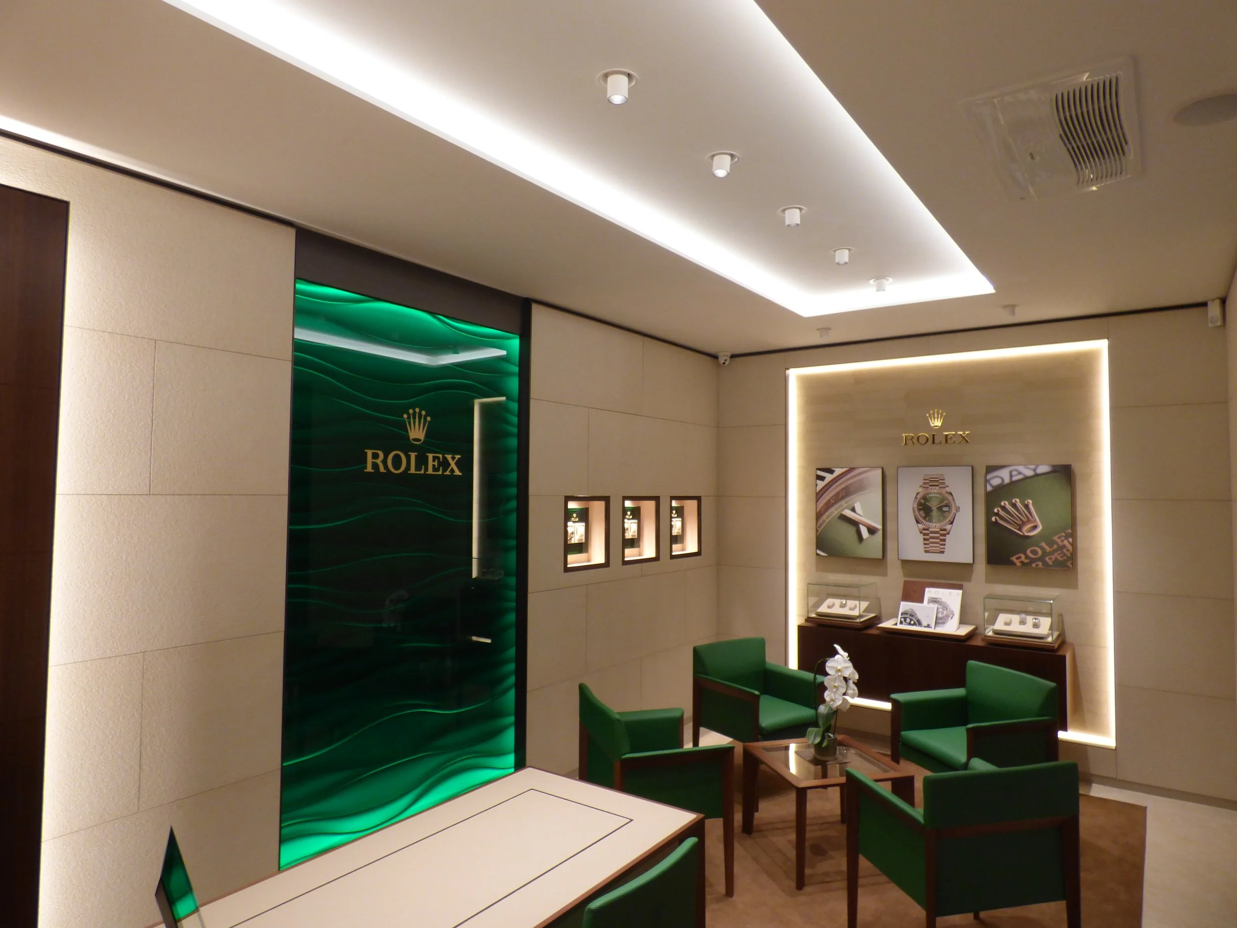 Innenaufnahme eines Rolex-Geschäfts mit grünen Stühlen, Vitrinen und Uhrenpräsentationen an der Wand, beleuchtete Schilder mit Rolex-Logos und Uhrenabbildungen.