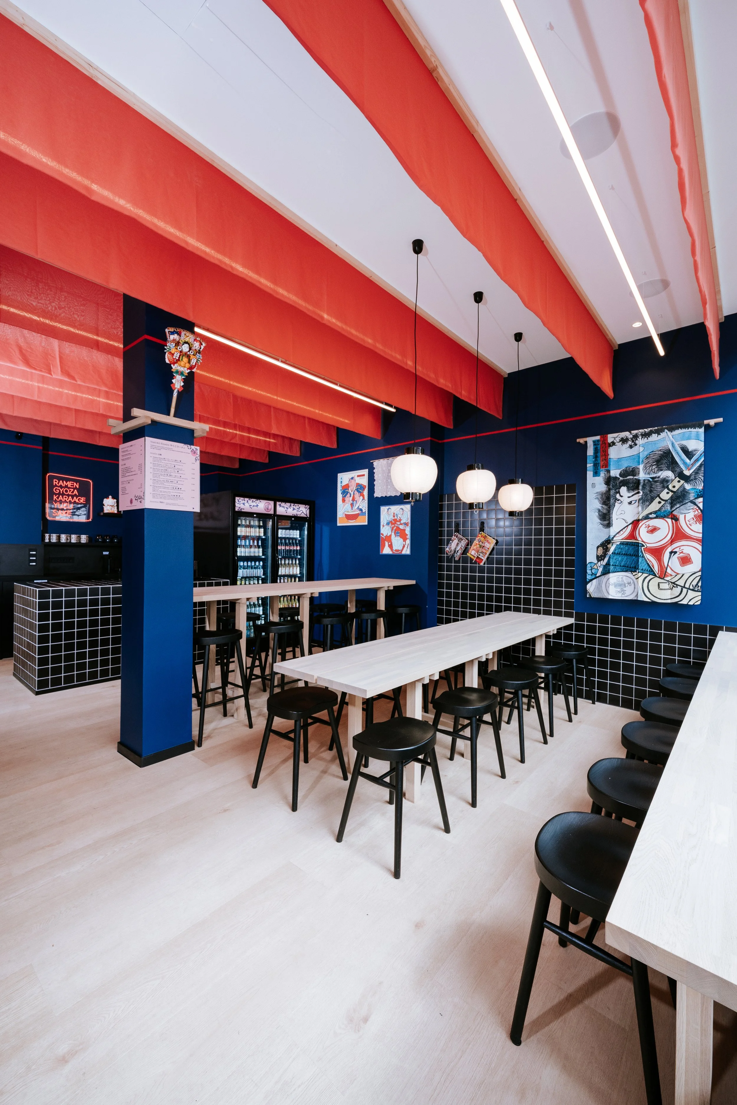 Innenansicht eines modernen Restaurants mit blauer Wandfarbe, roten Stoffvorhängen an der Decke, Taiko-Kunst an der Wand und Barhockern vor einem weißen Tisch.