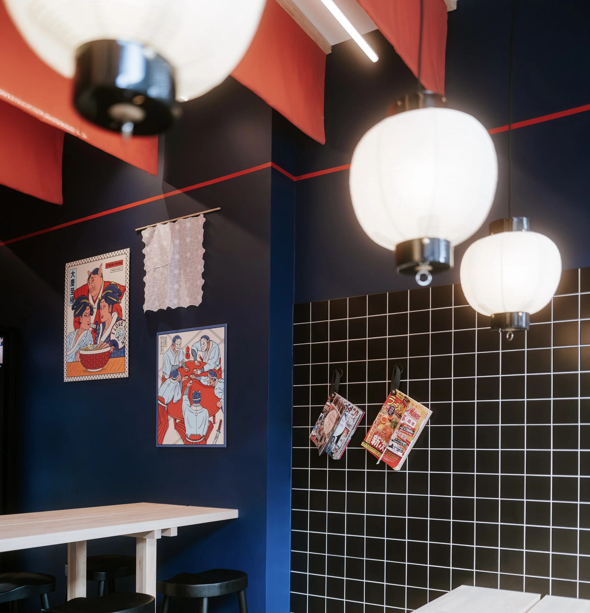 Innenaufnahme eines Zimmers mit dunkelblauer Wand, rotem Akzent, schwarzen und weißen Lampen, und japanischer Wanddekoration, darunter Poster mit japanischen Themen, sowie Magazine am Wandregal.