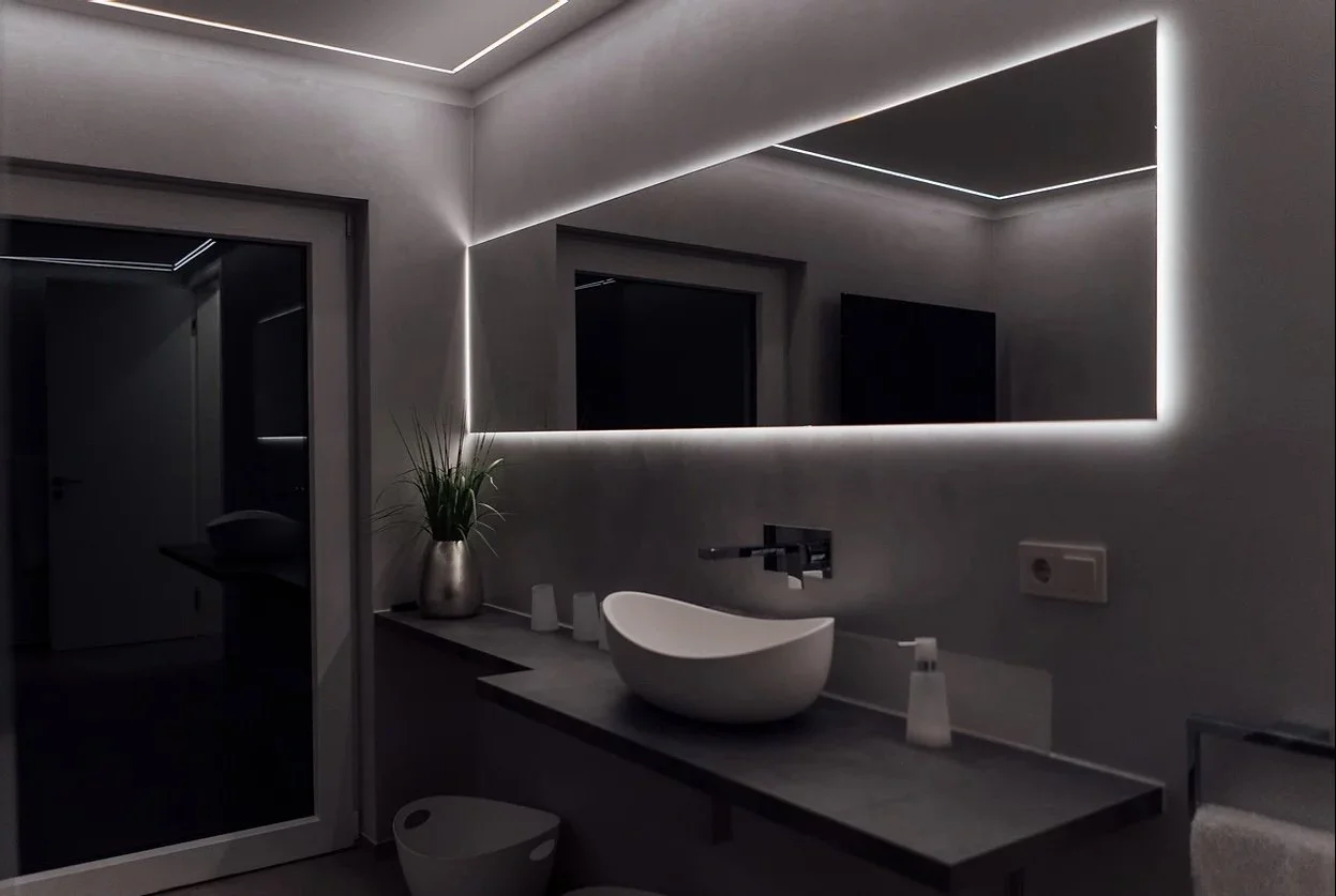Moderner Badezimmer mit LED-beleuchtetem Wandspiegel, Waschbecken, Pflanze im Metalltopf und dunklen Türen.