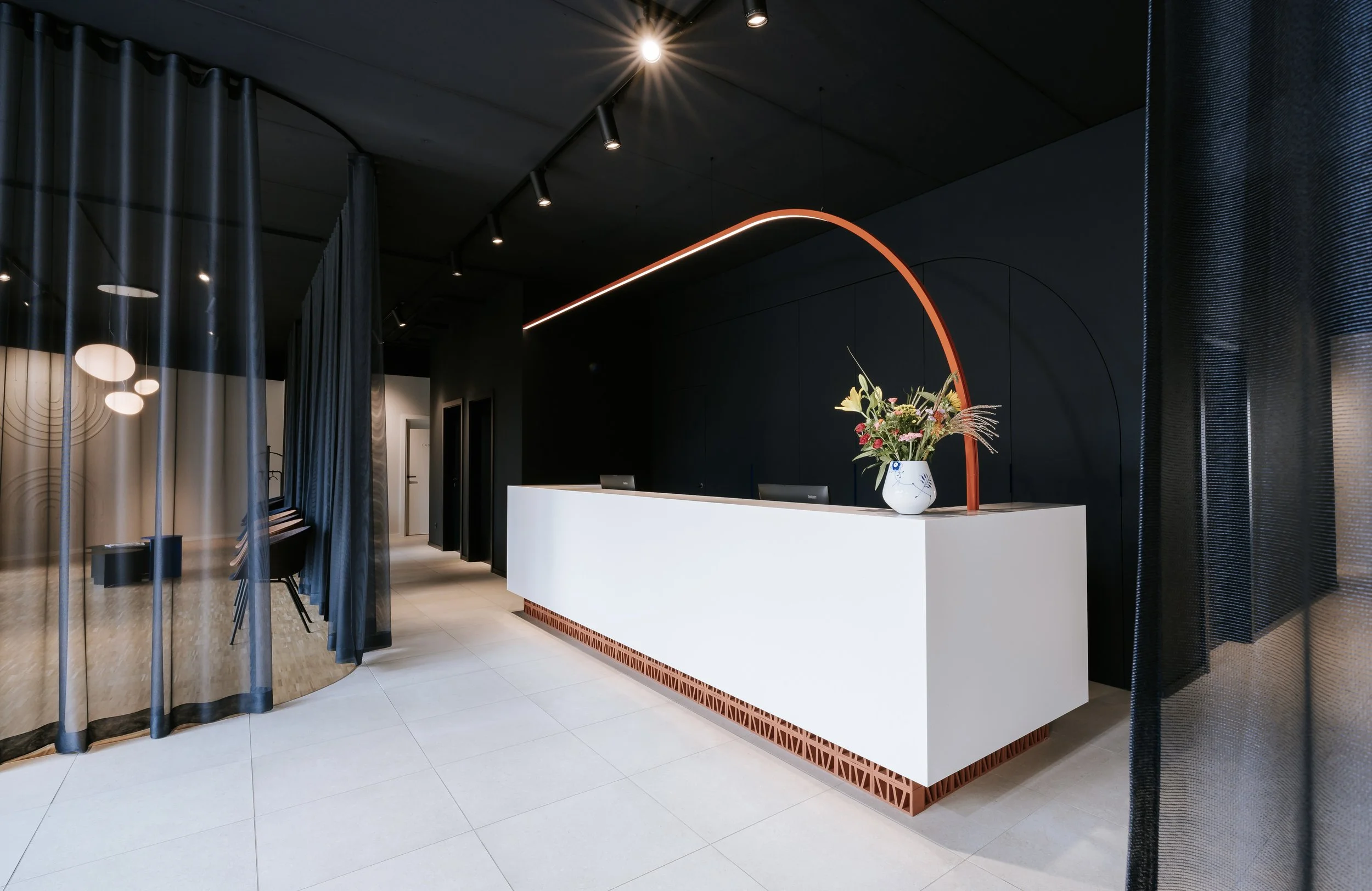 Moderne Augenklinik mit weißer Rezeption, bunter Blumenstrauß in einer weißen Vase, dunklen Wänden und einem roten Bogen. Hängende Lampen und Vorhänge im Vordergrund.
