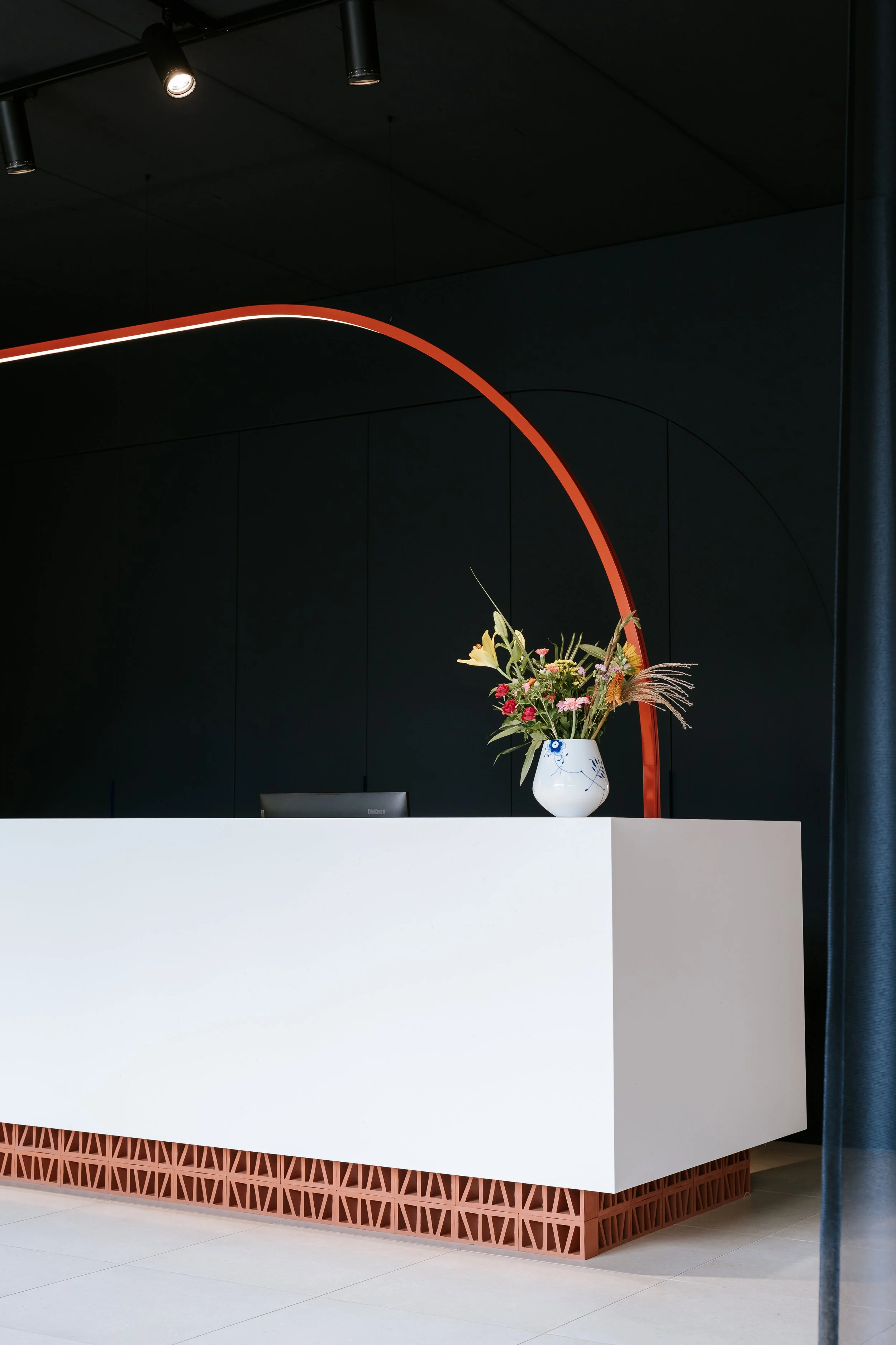 Ein Blumenstrauß in einer weißen Vase auf einer weißen Empfangstheke, im Hintergrund eine dunkle Wand und ein gebogener roter Rahmen, helle Bodenfliesen.