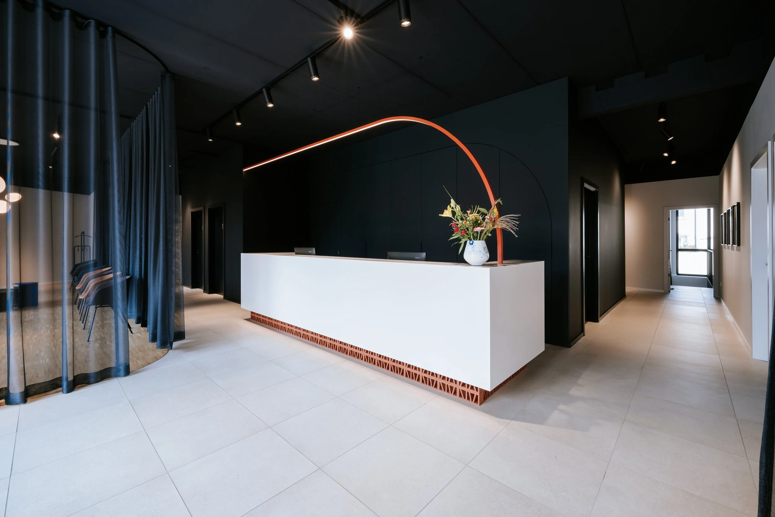 Moderner Empfangsbereich mit weißen Empfangstresen, Blumenstrauß in einer weißen Vase, dunkler Wand im Hintergrund, helle Fliesen, rollbarer Vorhang auf der linken Seite, Fenster und Türen im Raum. 