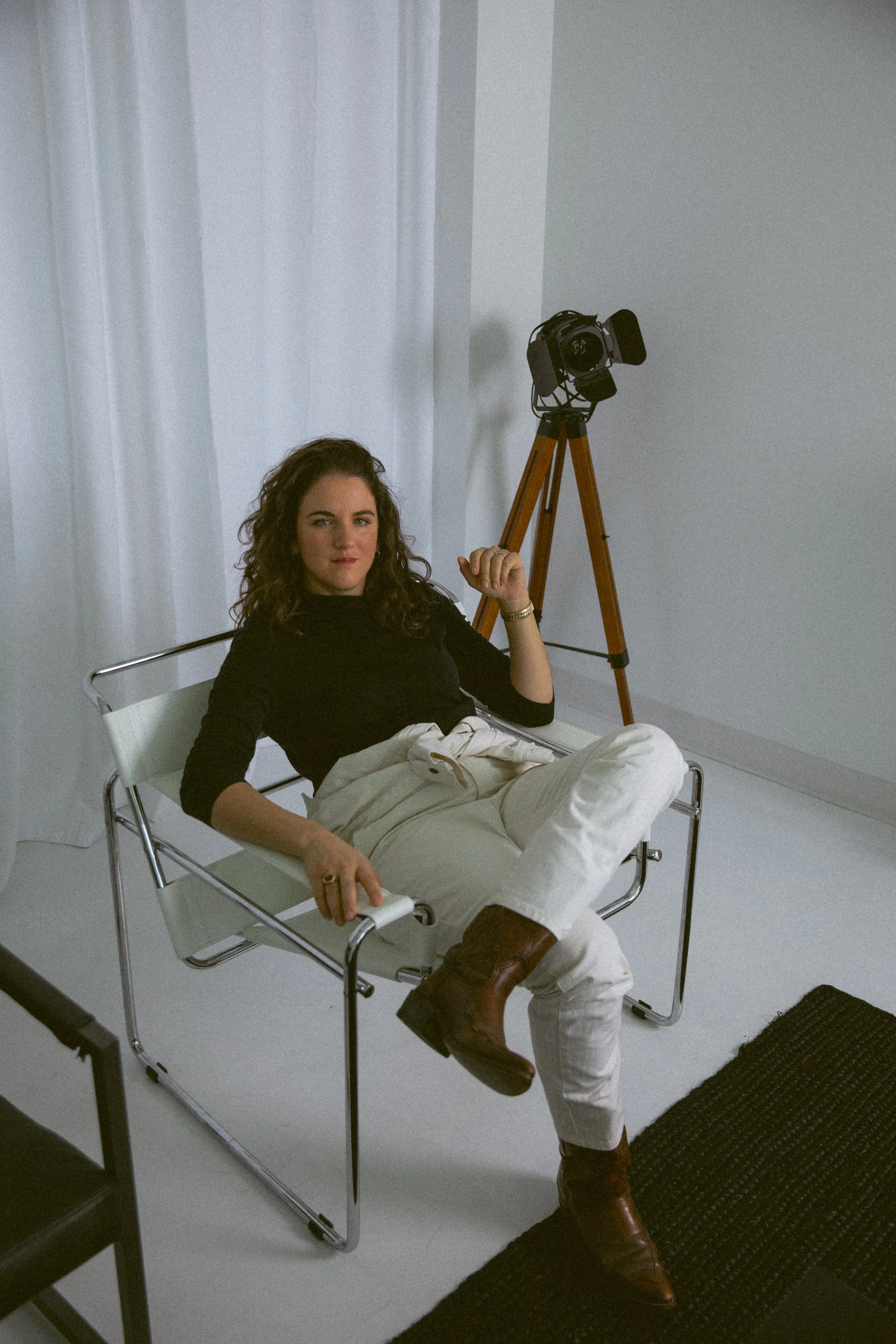 A woman with dark curly hair sitting on a white lounge chair, wearing beige pants, a black top, and brown boots, holding a remote control. She is in a room with white walls, a white curtain, a black rug, and a vintage camera on a tripod in the background.