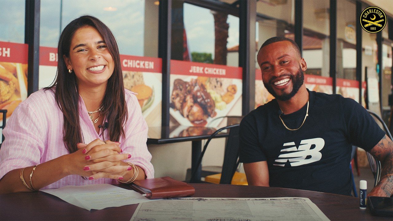 Two people sitting at a table in a restaurant, smiling at the camera. The woman on the left has long brown hair, wears a pink striped shirt, and the man on the right has a beard, short hair, and wears a black t-shirt with a white logo. The restaurant has menu boards with images of chicken dishes in the background.