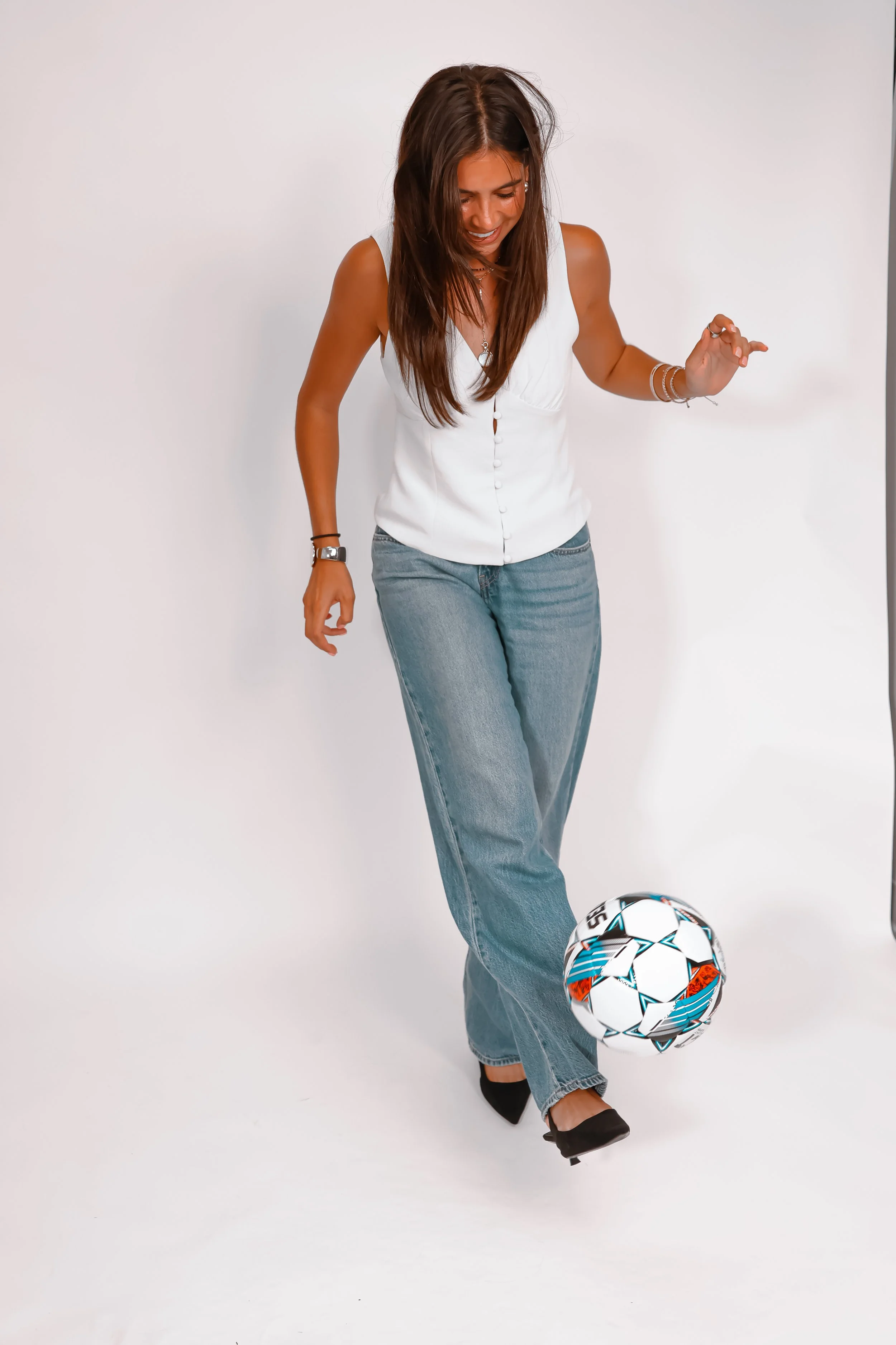 A woman with long brown hair, wearing a white sleeveless top, blue jeans, and black shoes, is standing against a plain white background. She is standing on one leg, with the other foot on a soccer ball, and appears to be playing or balancing with it.
