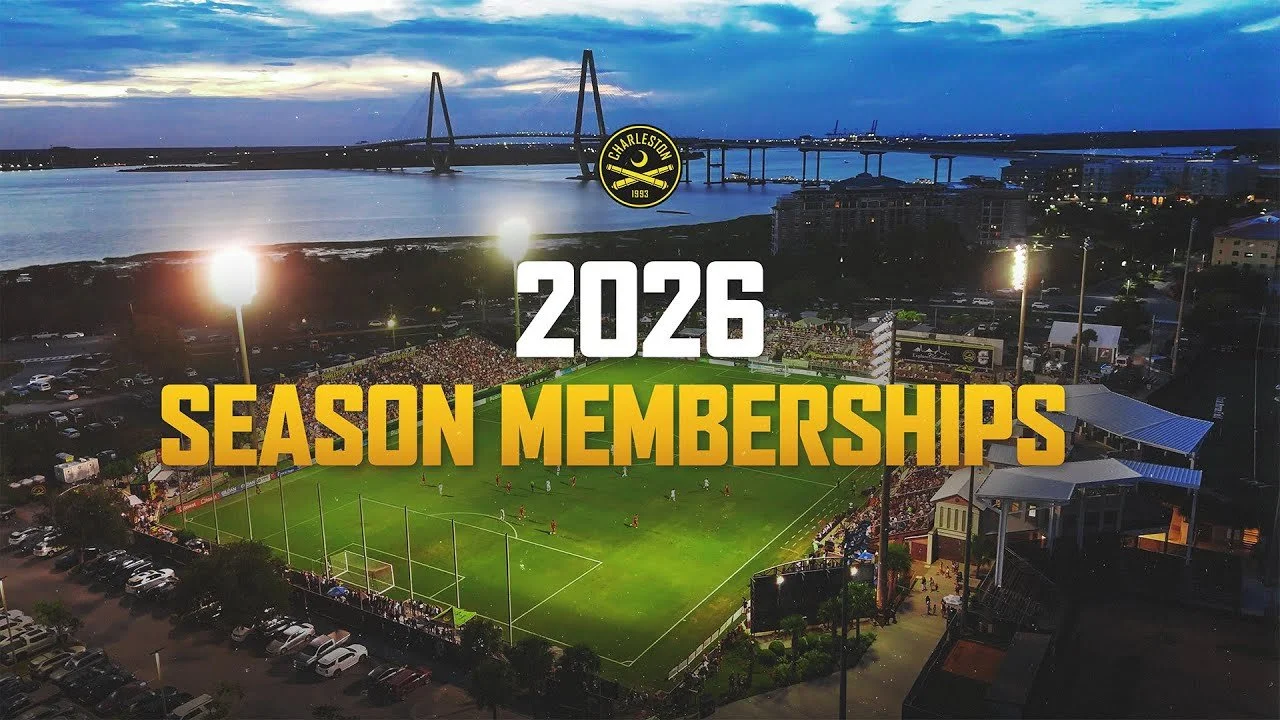 Aerial view of a football stadium with a crowd during twilight, showing a game in progress, parking lot adjacent, and a river in the background with a bridge, overcast sky, and printed text announcing Charleston 2026 season memberships.