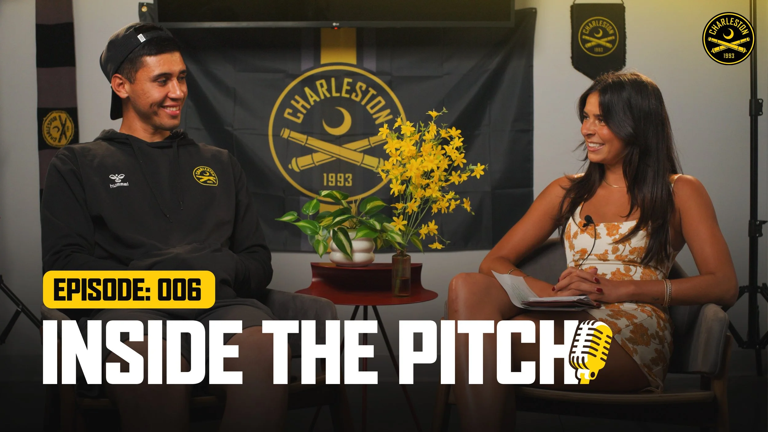 Two people engaged in a conversation during a podcast recording, with Charlestion soccer team banners and logos in the background, including a large yellow flower arrangement on the table.