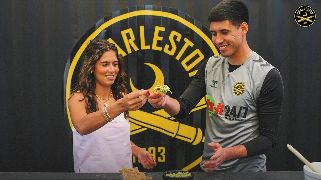 A woman and a man smiling as the woman receives a piece of food from the man in front of a Charleston team logo.